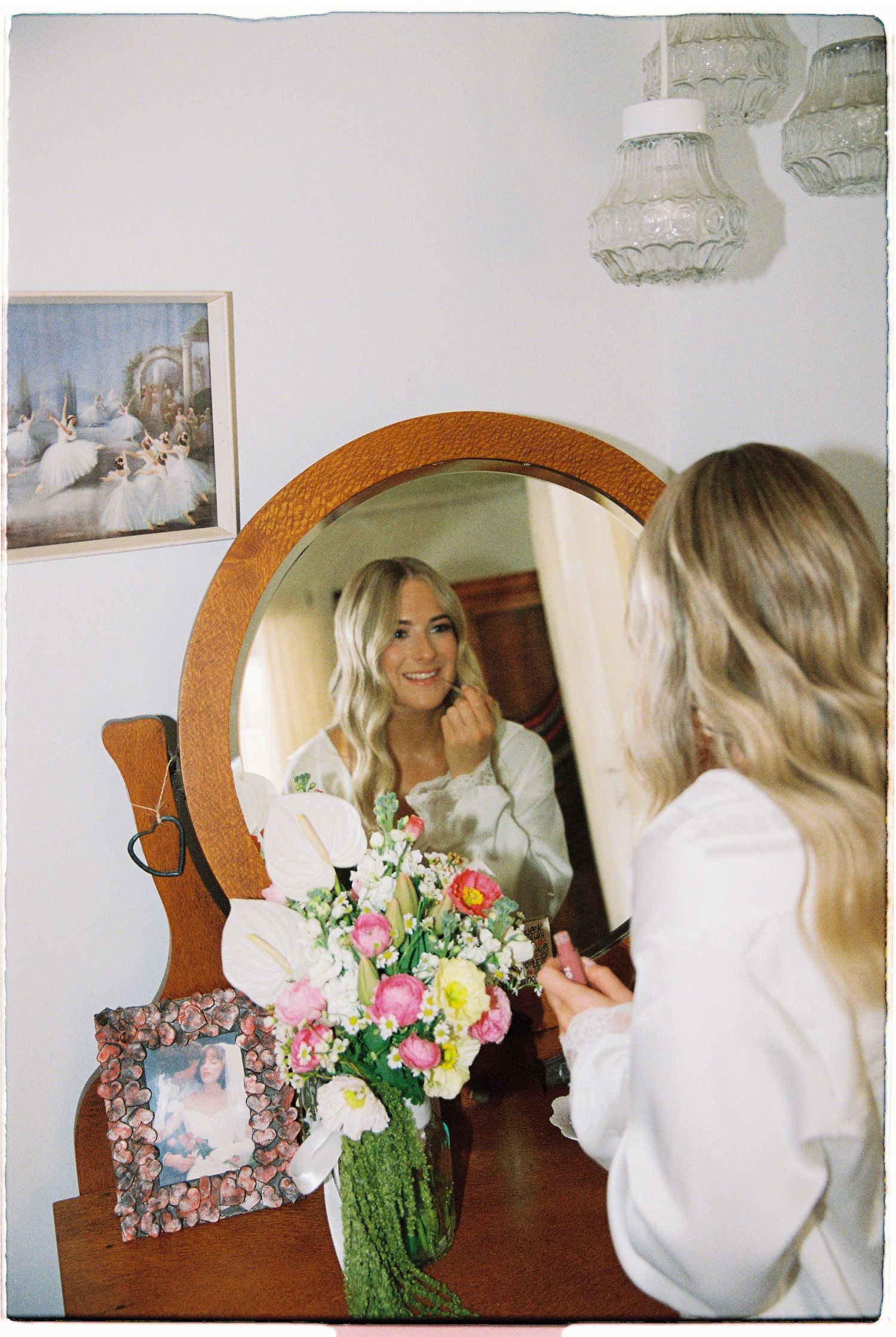 Blonde bride in white robe admires her reflection in a round wooden mirror beside a colorful floral bouquet.