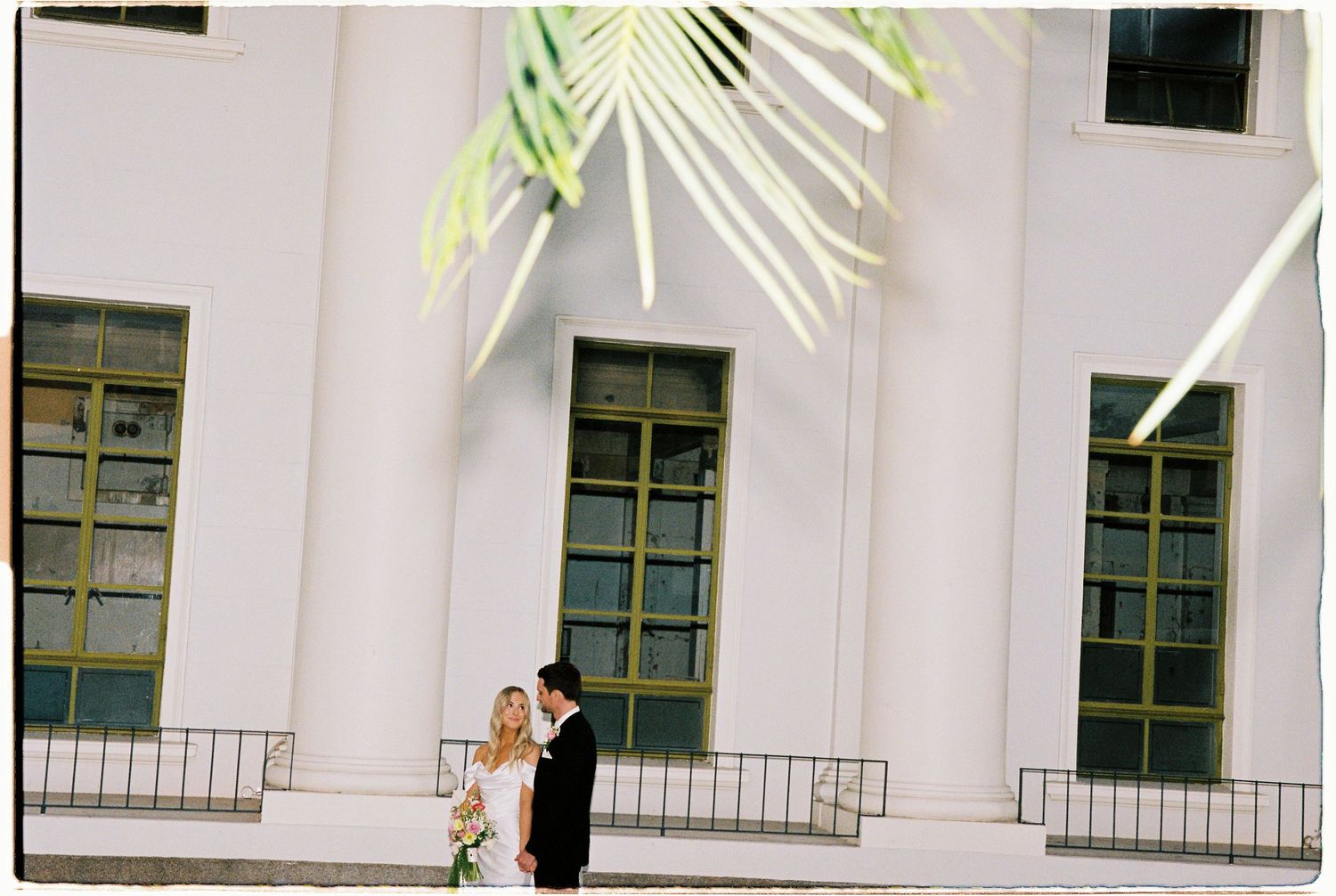 Wedding couple sharing a romantic moment outside a white building with palm frond shadows overhead.