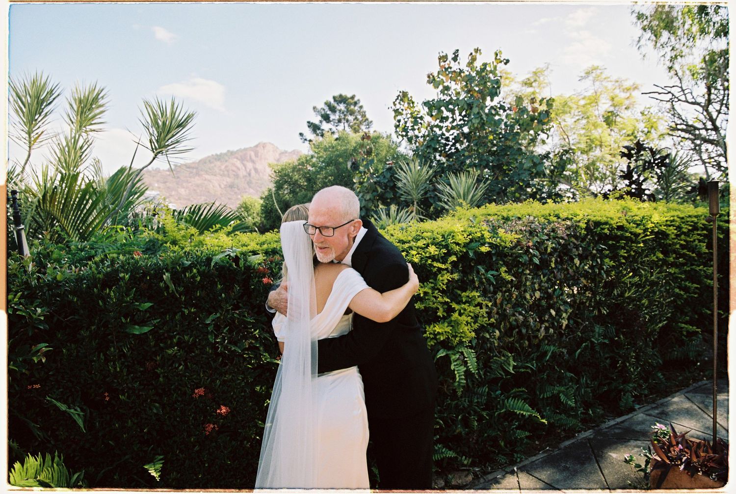 Emotional wedding guest embracing bride in white veil against lush green hedge with mountain backdrop.