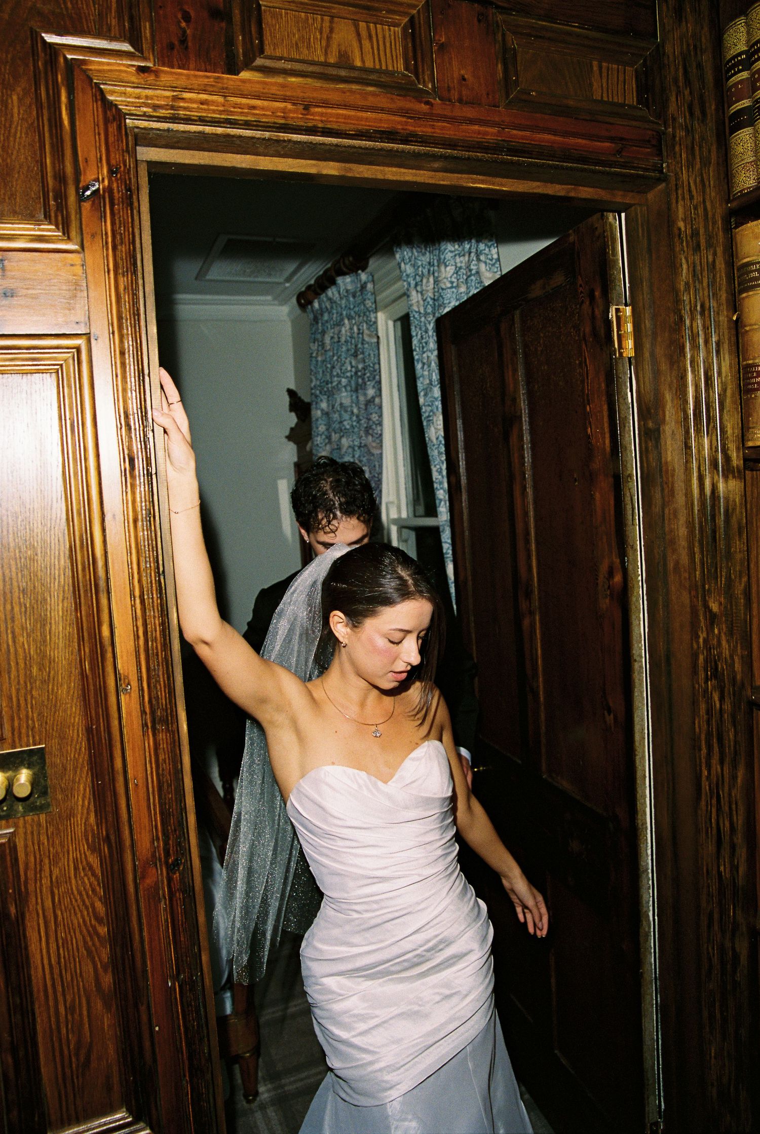 Bride in white strapless gown and veil walking through ornate wooden doorway with groom behind her.