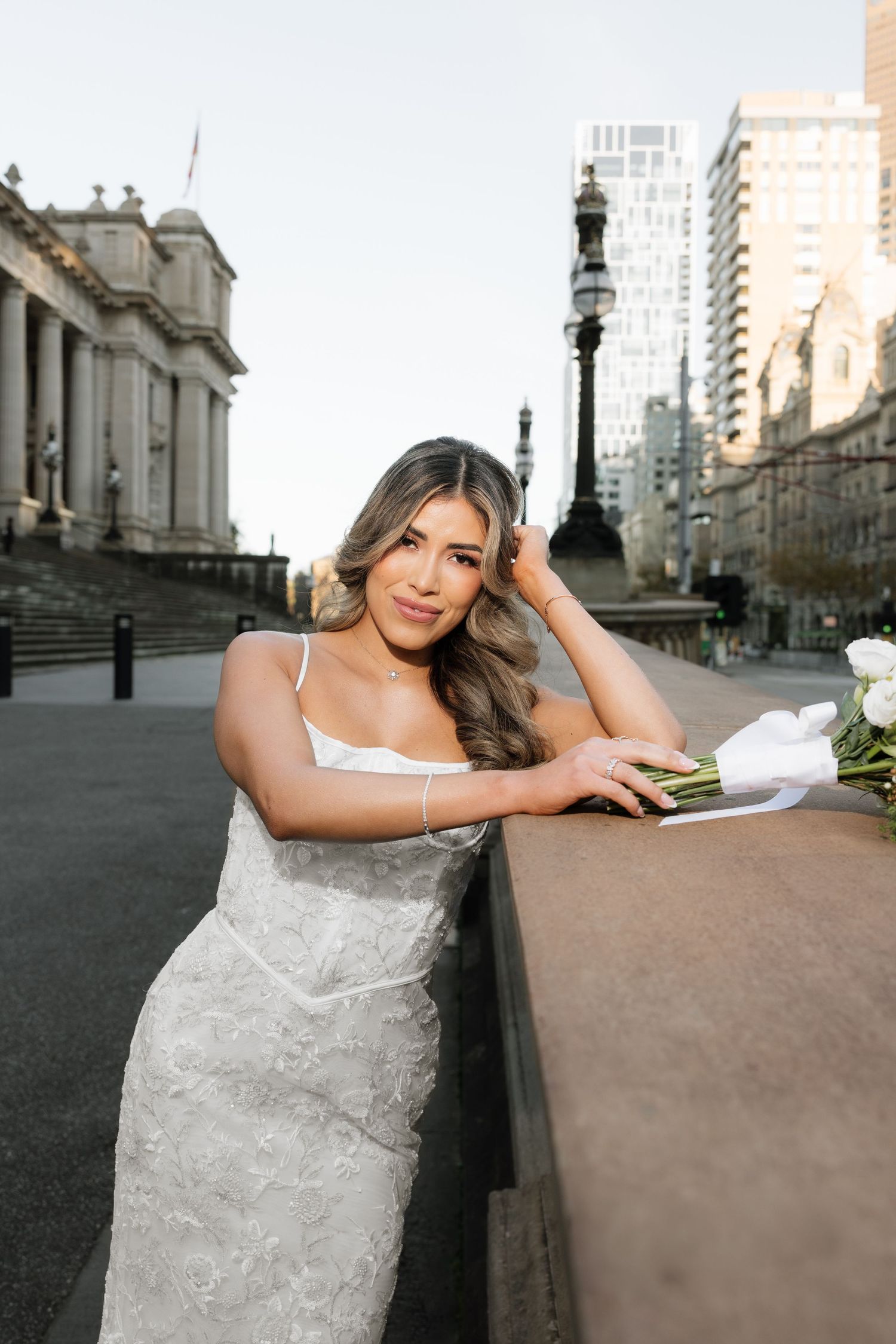 Bride in white lace gown holding bouquet, leaning on stone wall with city buildings backdrop.