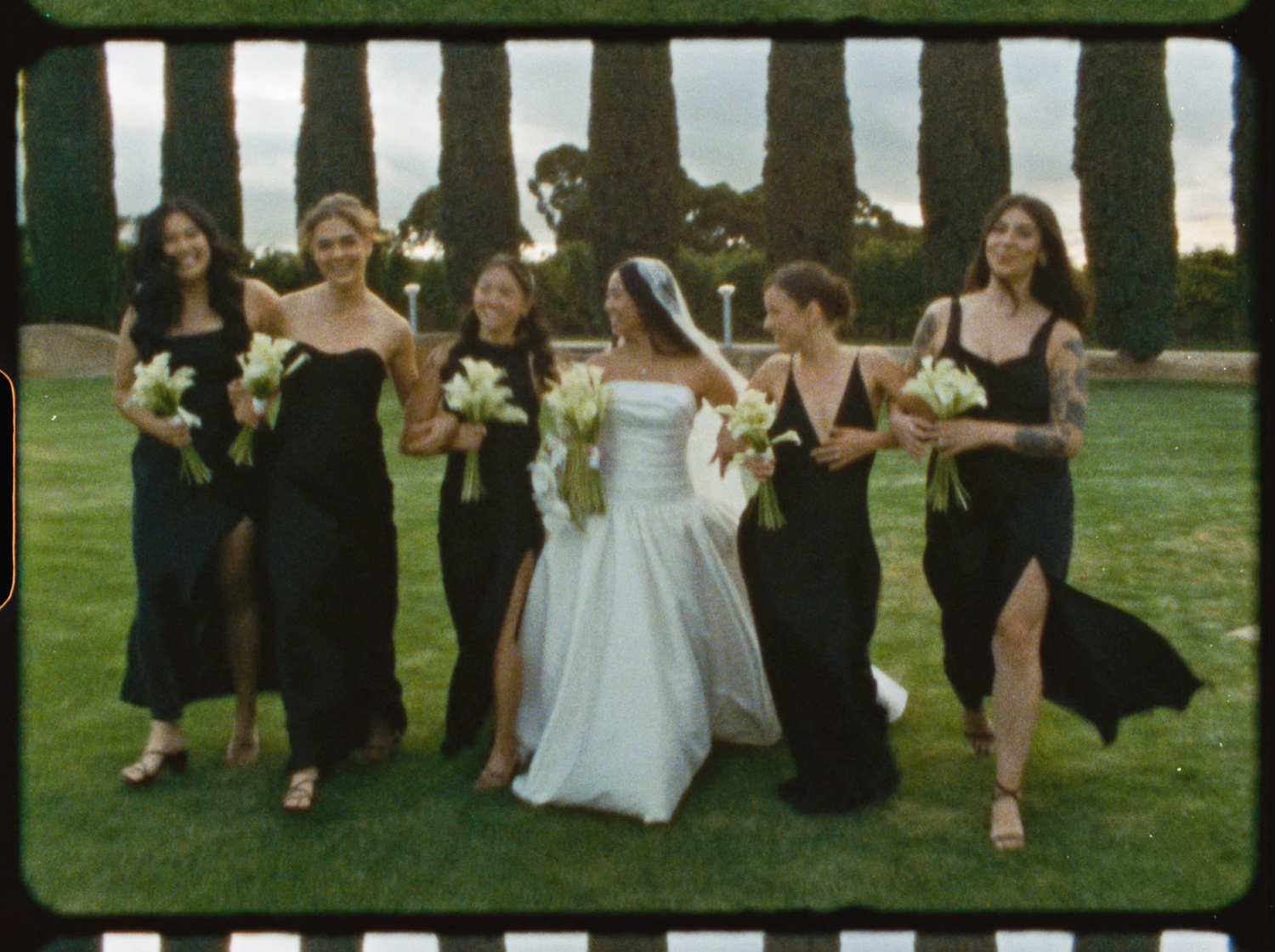 Bride in white gown walks with six bridesmaids in black dresses holding calla lilies outdoors.