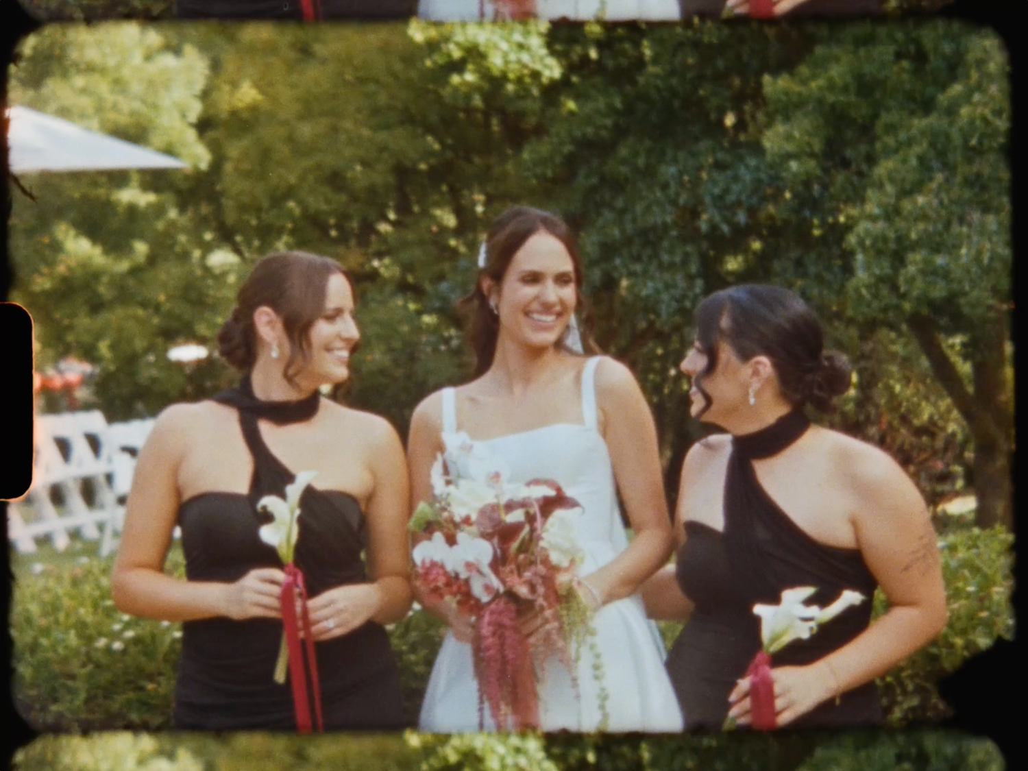 Smiling bride in white dress flanked by two bridesmaids in black dresses at garden wedding.