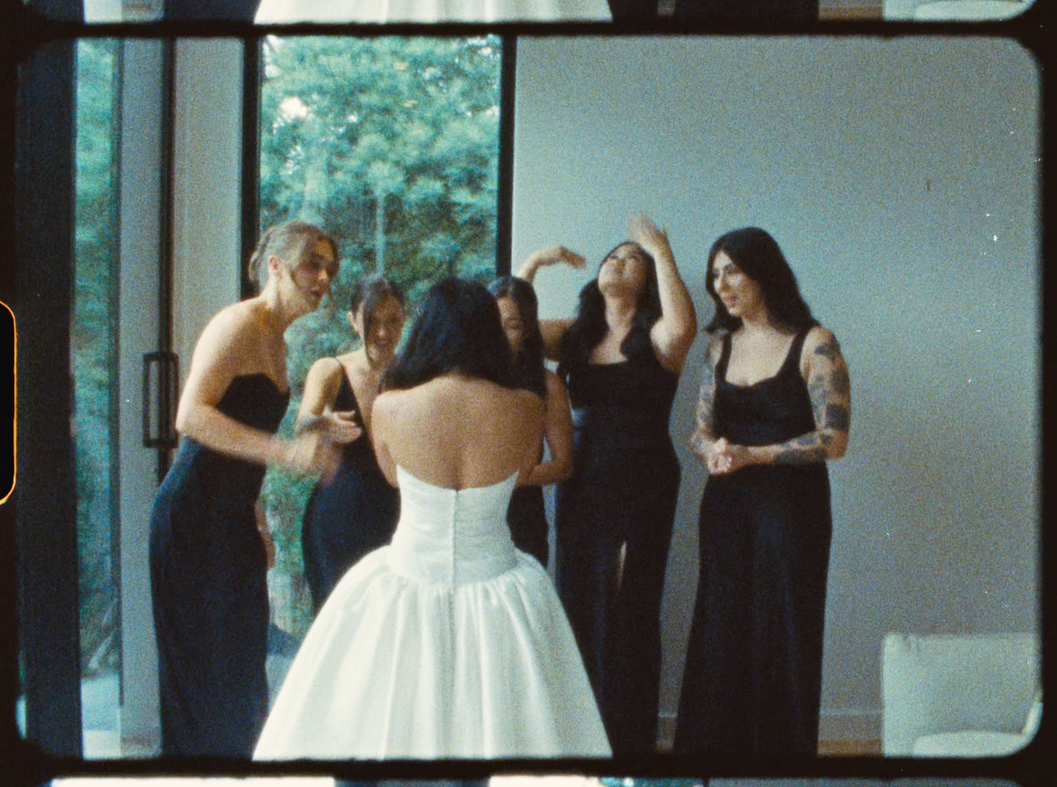 Bridesmaids in black dresses form a heart gesture for the bride in a white gown by large windows.