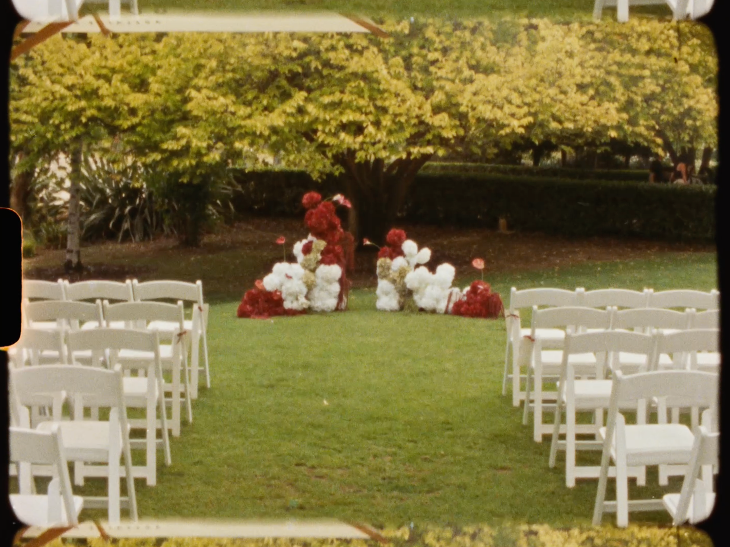 Outdoor wedding ceremony setup with white chairs and red and white balloon arch on a lush green lawn.