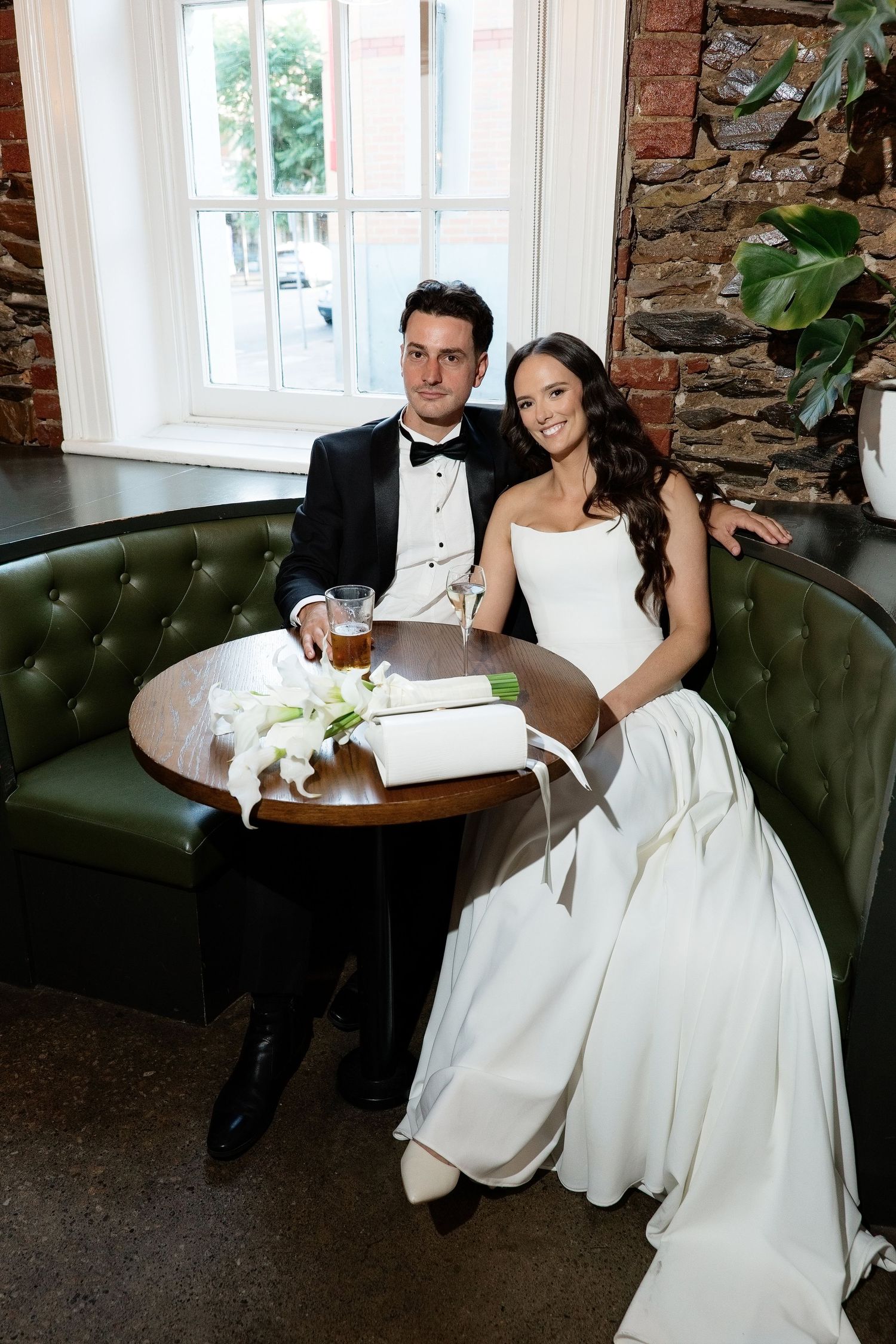 Wedding couple posing at a round table on a green leather sofa near a sunlit window in a pub.