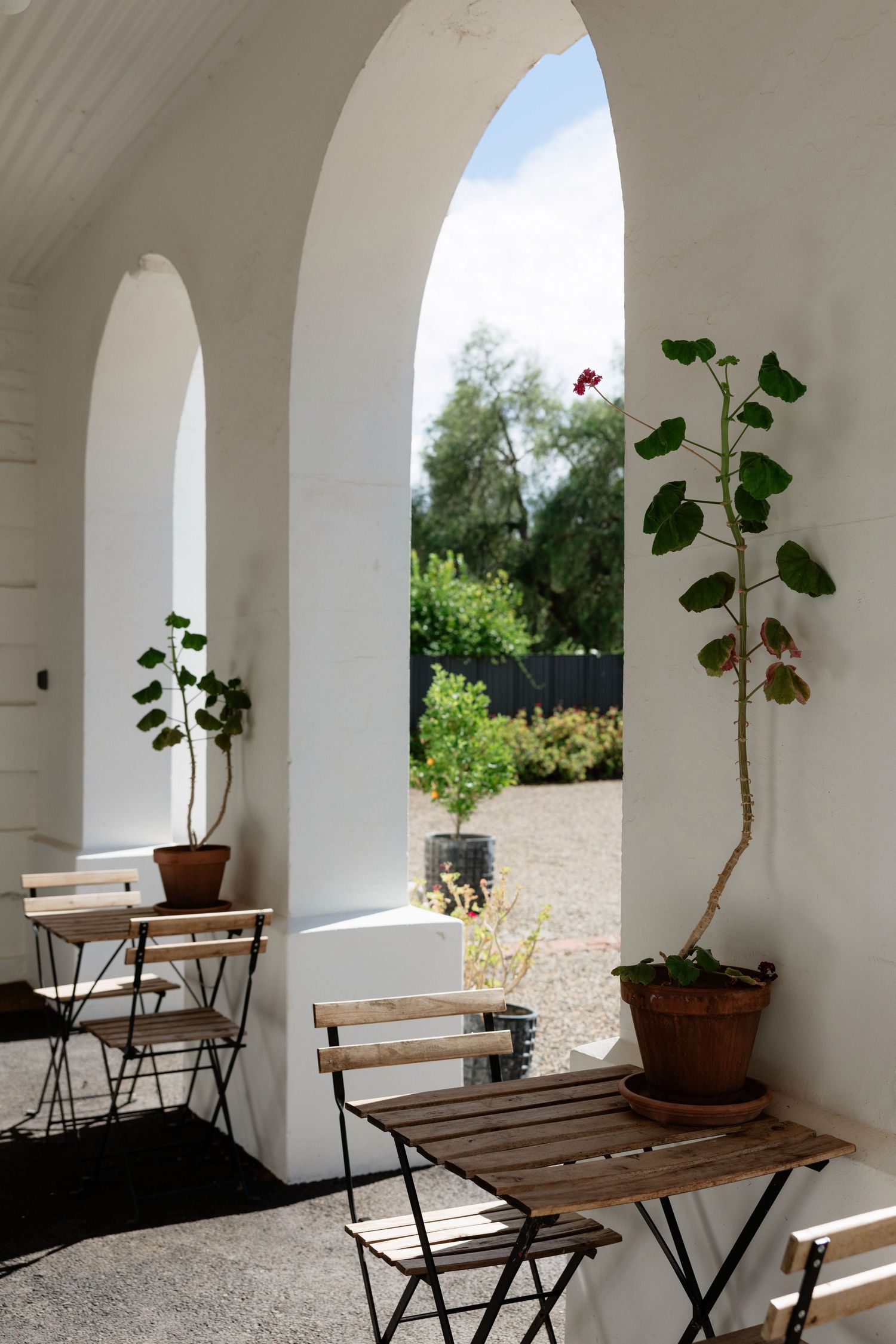 Sunlit white arched veranda with wooden bistro chairs, potted geraniums, and garden views beyond.
