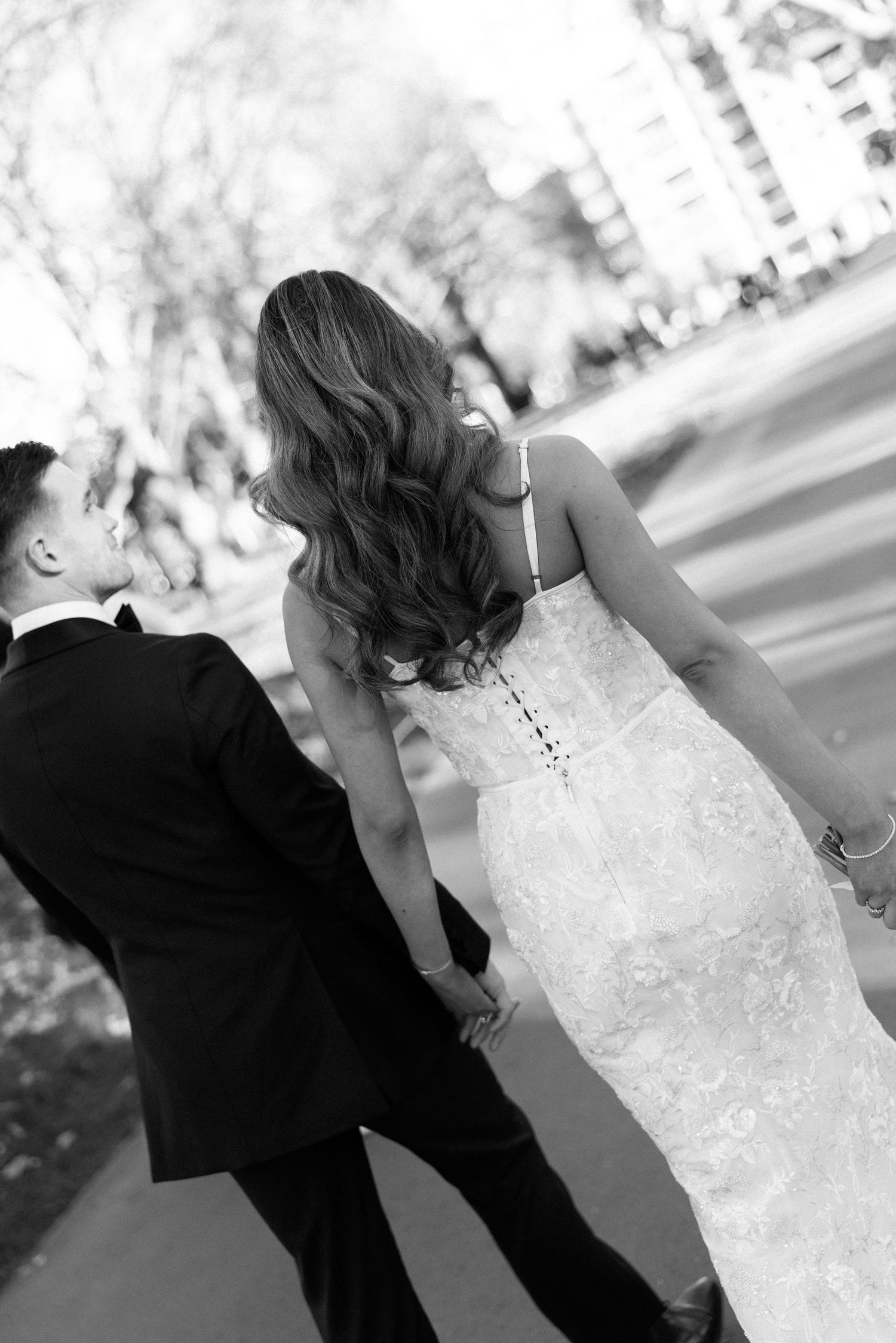 Black and white photo of a bride in lace gown and groom in tuxedo walking together outdoors.