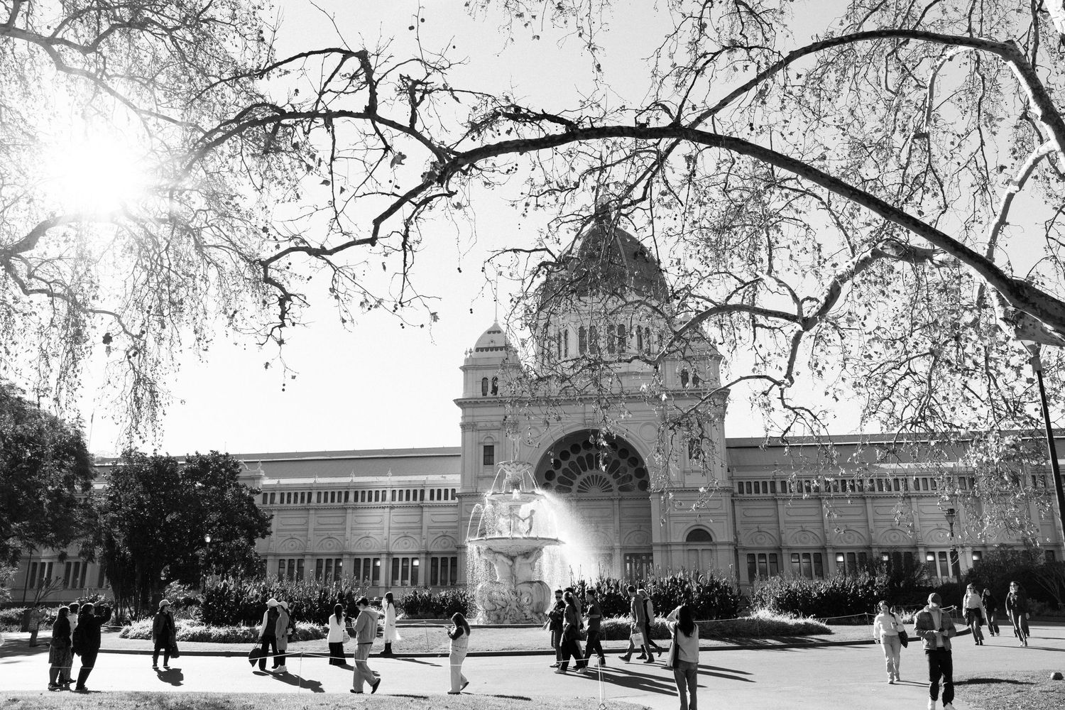 Black and white photo of the Royal Exhibition Building in Melbourne with visitors strolling through Carlton Gardens.