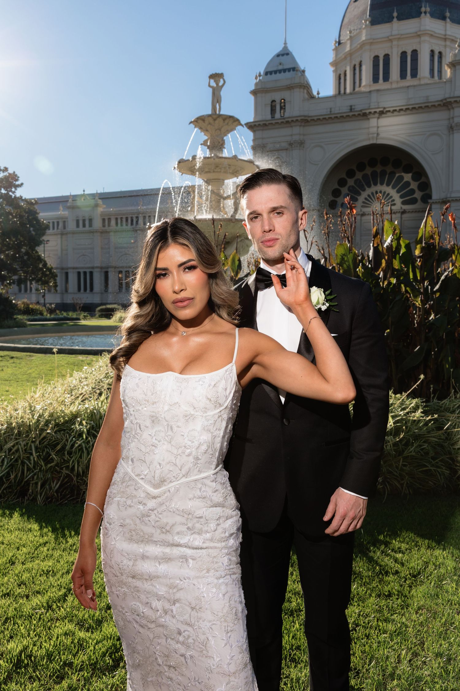 Bride in white lace gown and groom in black tuxedo pose before ornate building with fountain.