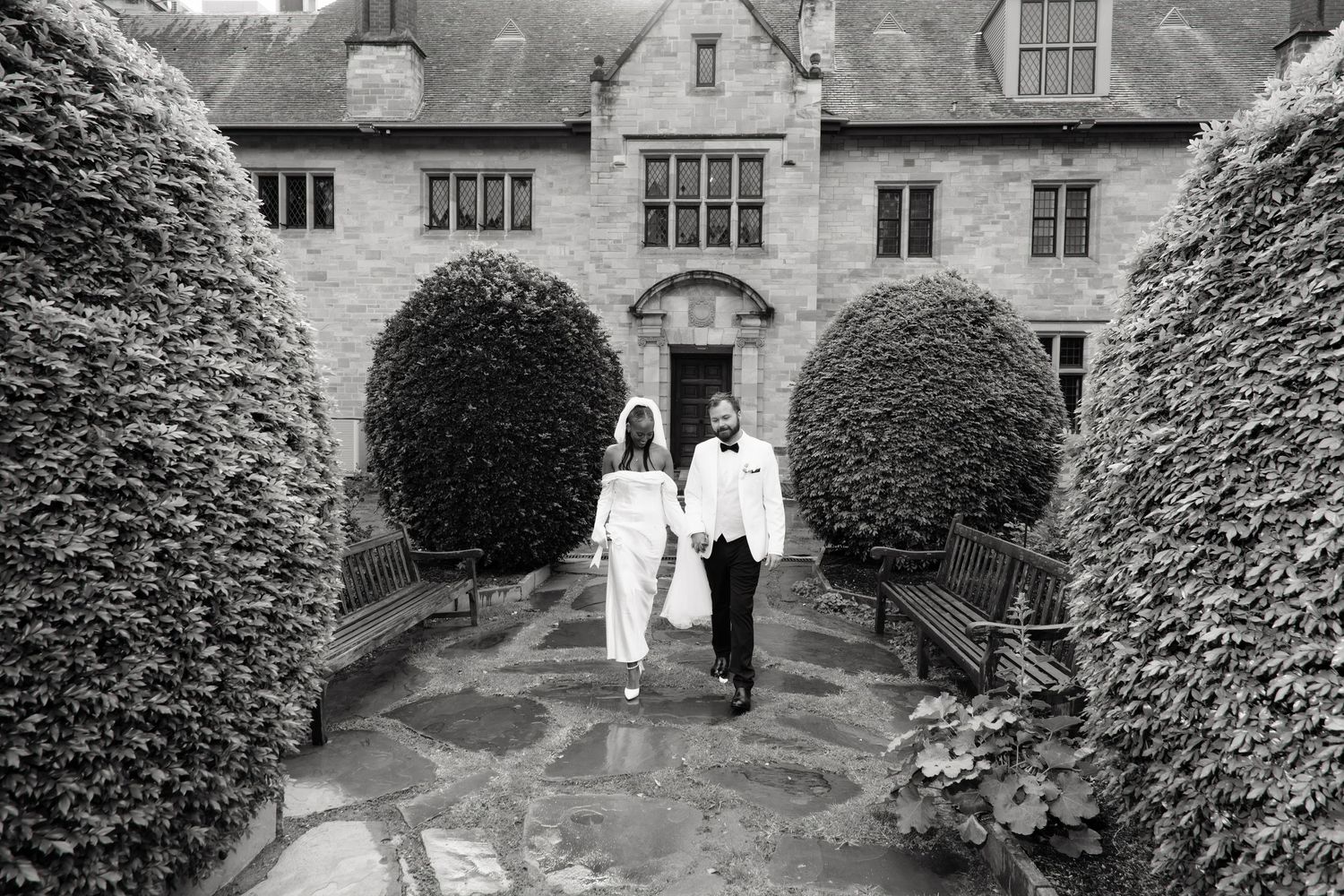 Black and white photo of bride and groom walking through manicured hedge garden of historic estate.