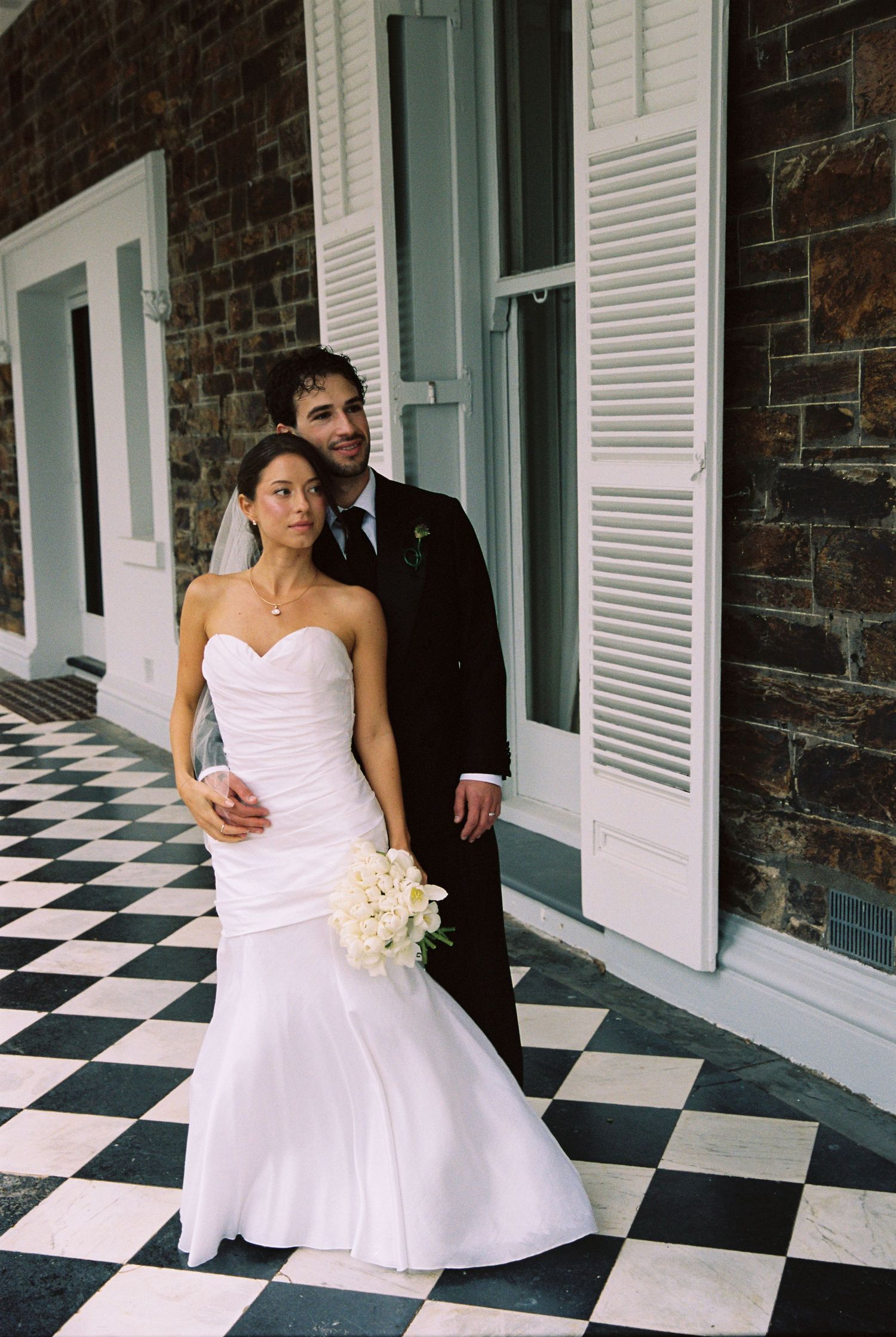 Bride and groom pose elegantly on black and white checkered floor beside historic brick building.
