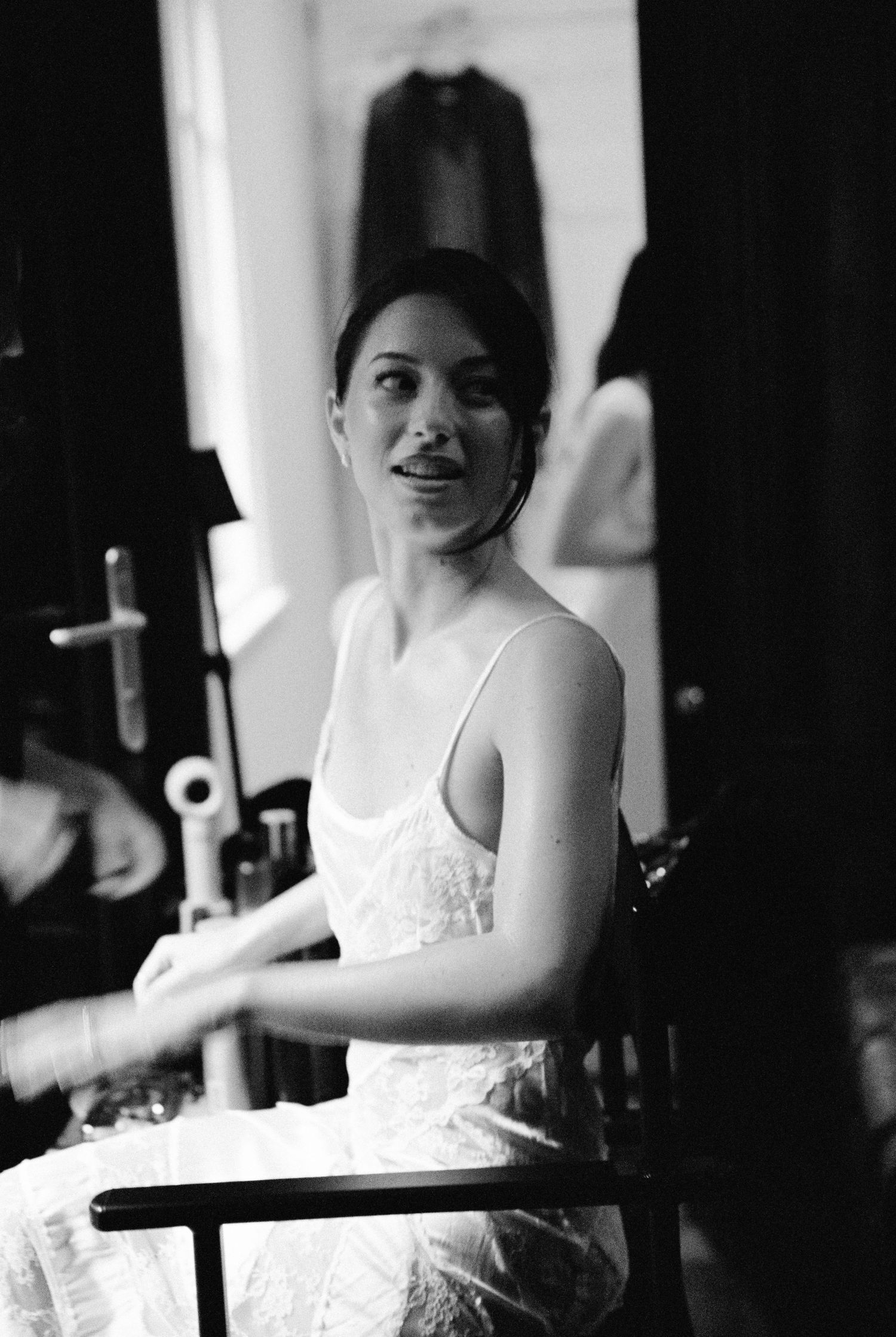Bride in white slip dress seated and smiling during wedding preparations in dressing room.