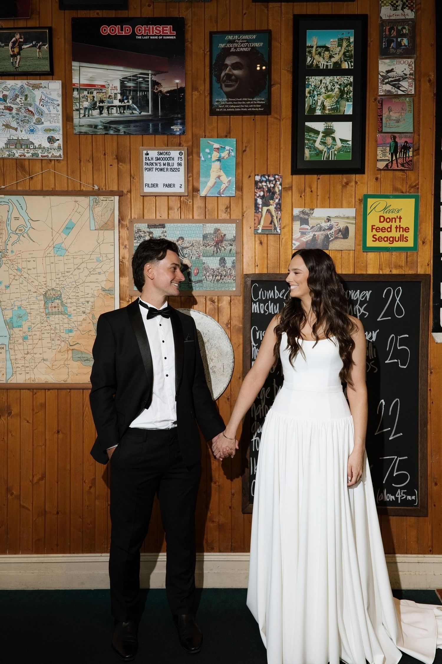 Bride in white gown and groom in tuxedo holding hands in front of a rustic wood-paneled wall.