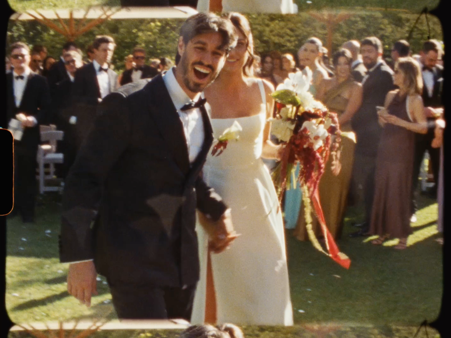 Laughing bride and groom walking through cheering wedding guests outdoors after garden ceremony.