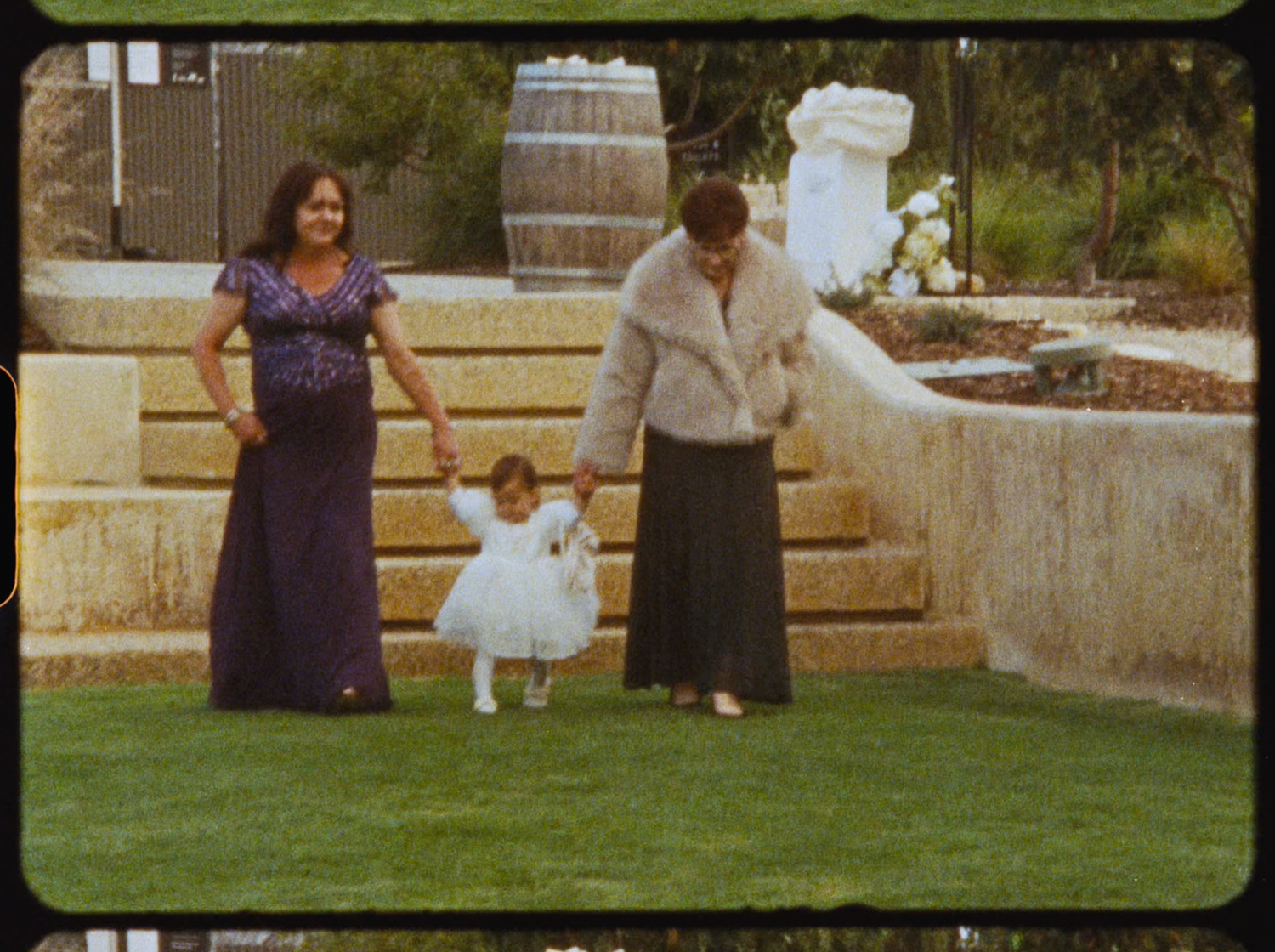Two women and a young girl in white dress posing on green lawn at a wedding venue.