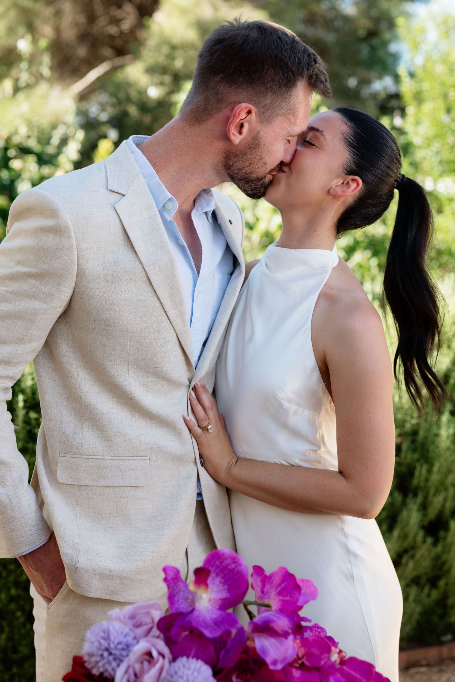 Bride and groom sharing first kiss with vibrant purple floral bouquet in foreground.