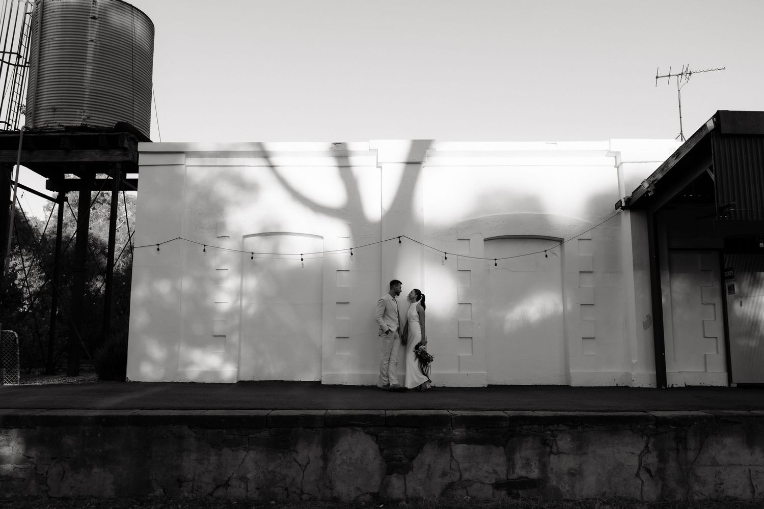 Bride and groom walking beside white wall with dramatic tree shadow, black and white wedding photo.