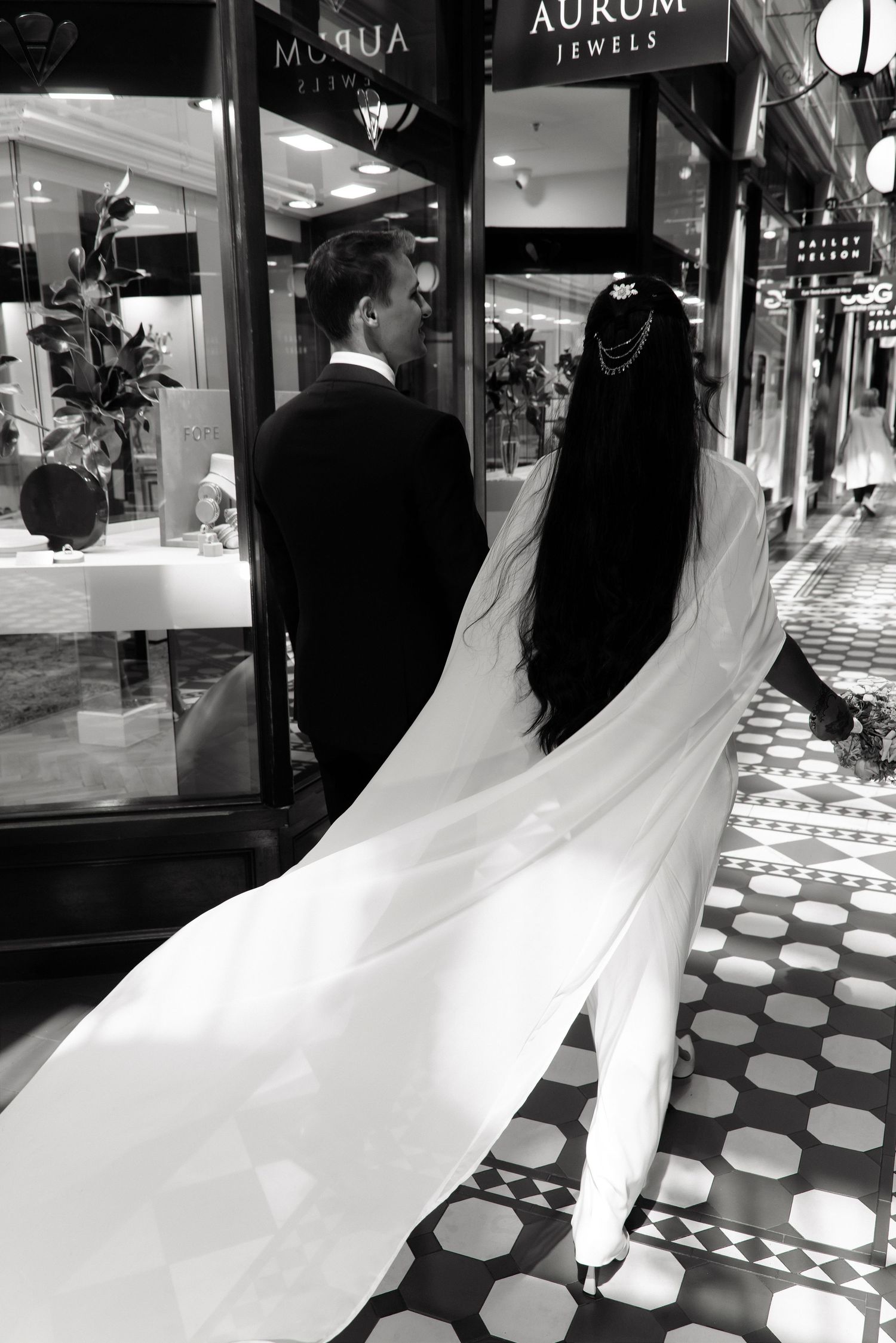 Black and white photo of bride and groom walking through elegant shopping arcade, veil flowing behind.