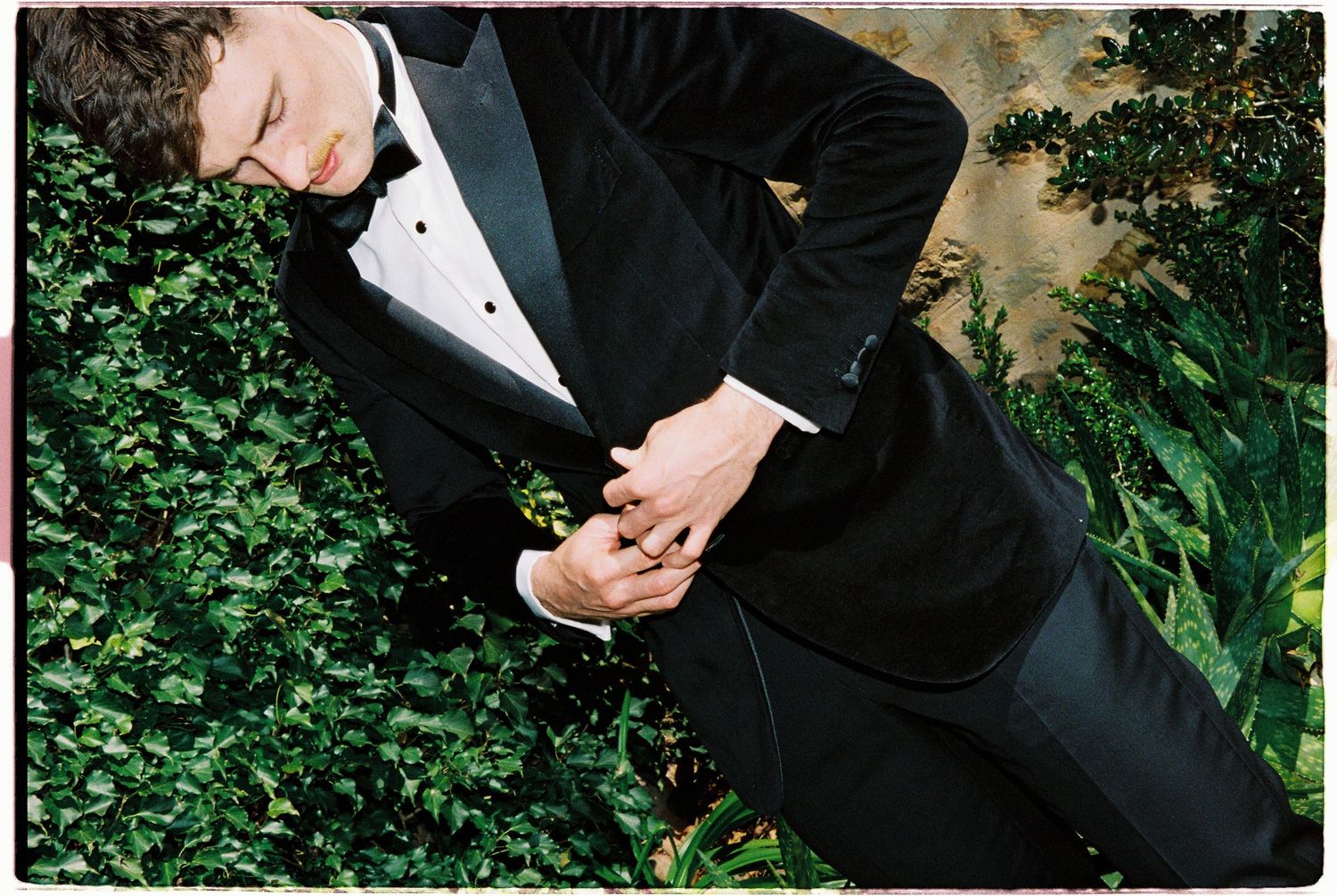 Groom in black tuxedo adjusting jacket against lush green hedge backdrop.