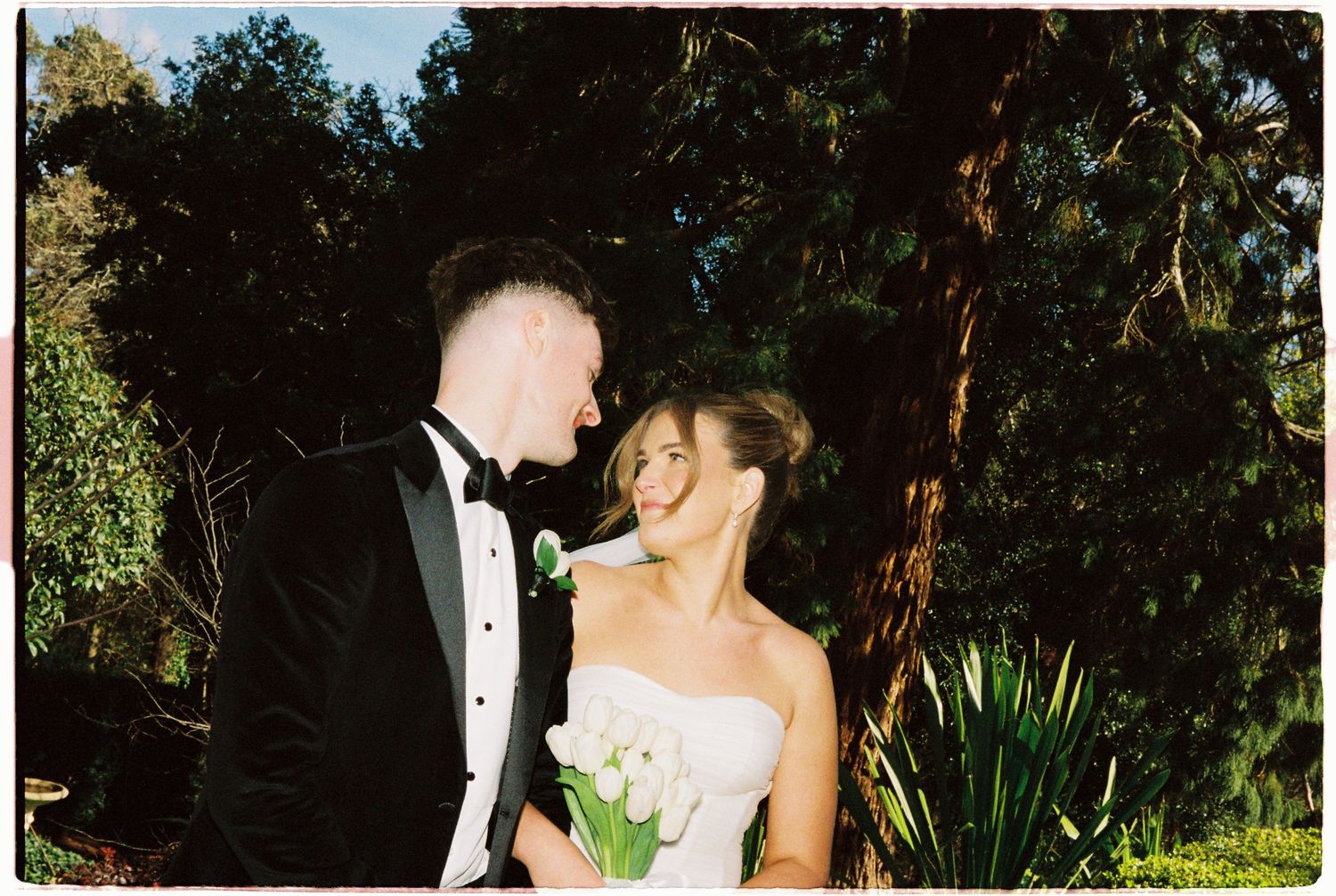 Smiling bride and groom in formal attire standing together in a garden.
