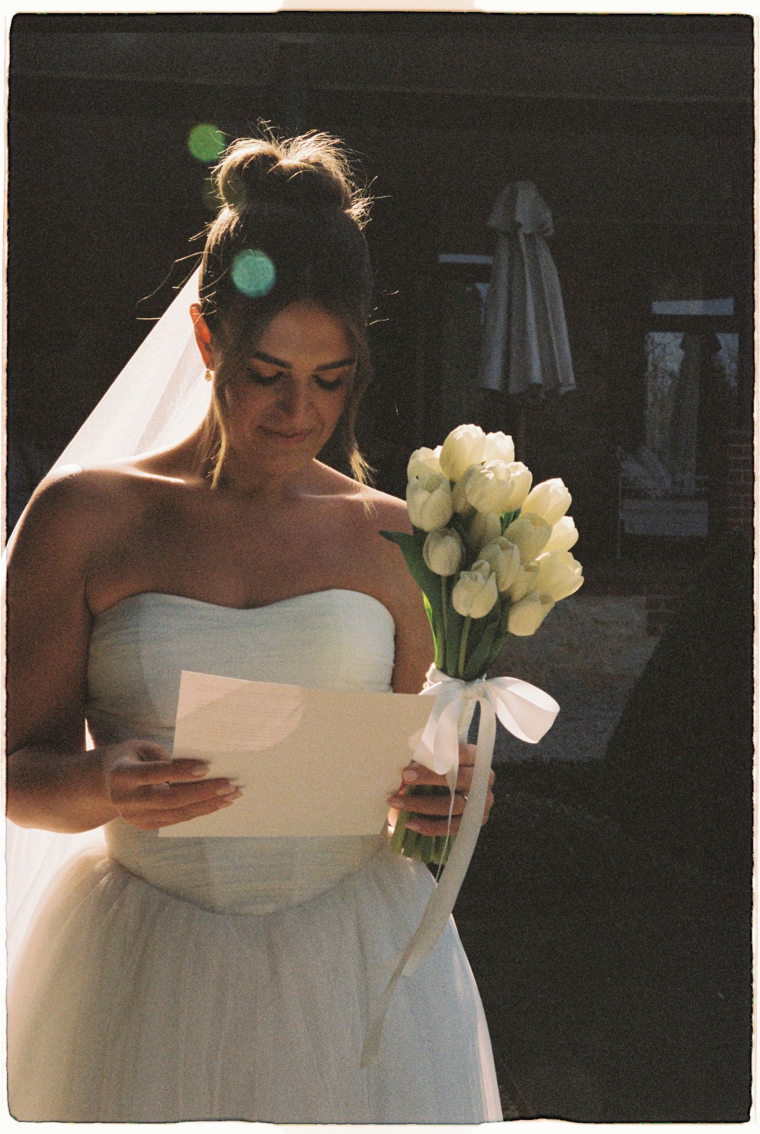 Bride in strapless white gown reads a letter while holding a white tulip bouquet in sunlight.
