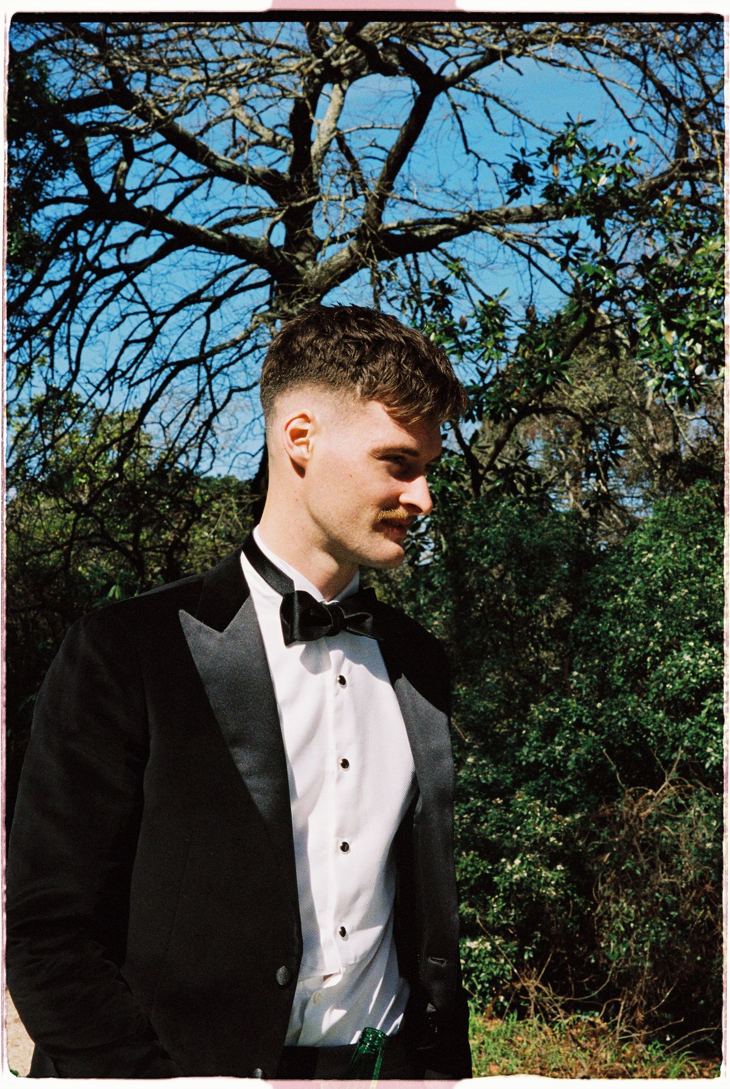 Young man in black tuxedo with bow tie standing outdoors against trees and blue sky.