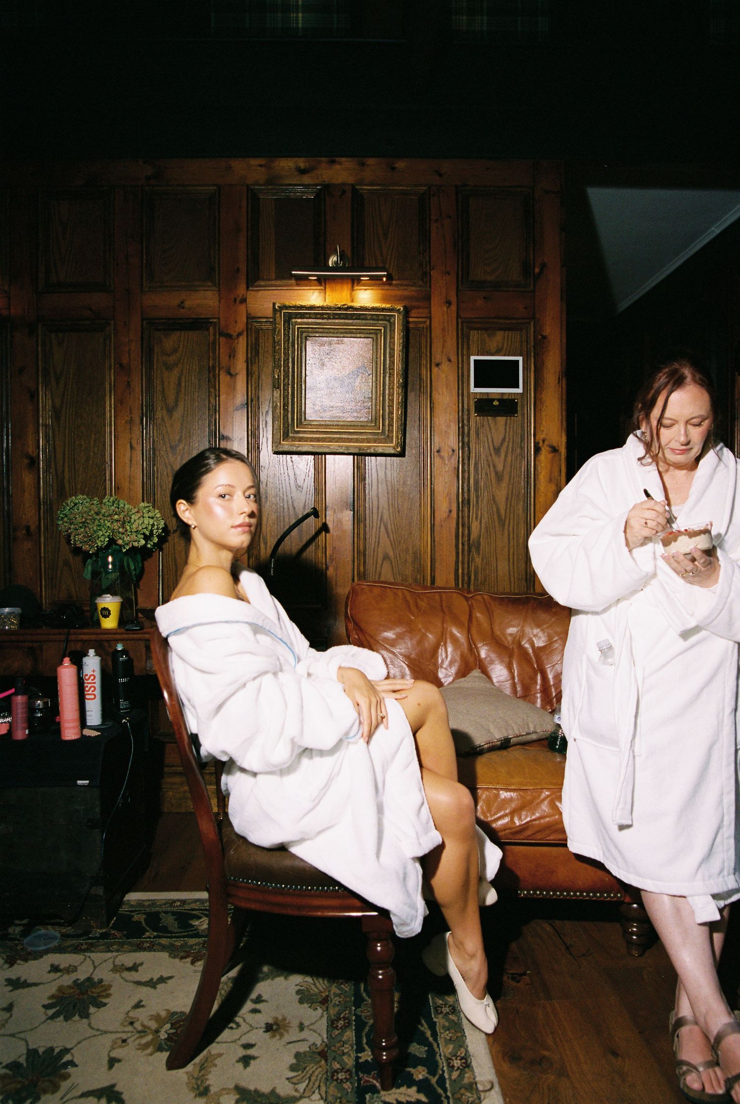 Bride and makeup artist in white robes preparing in elegant wood-paneled bridal suite.