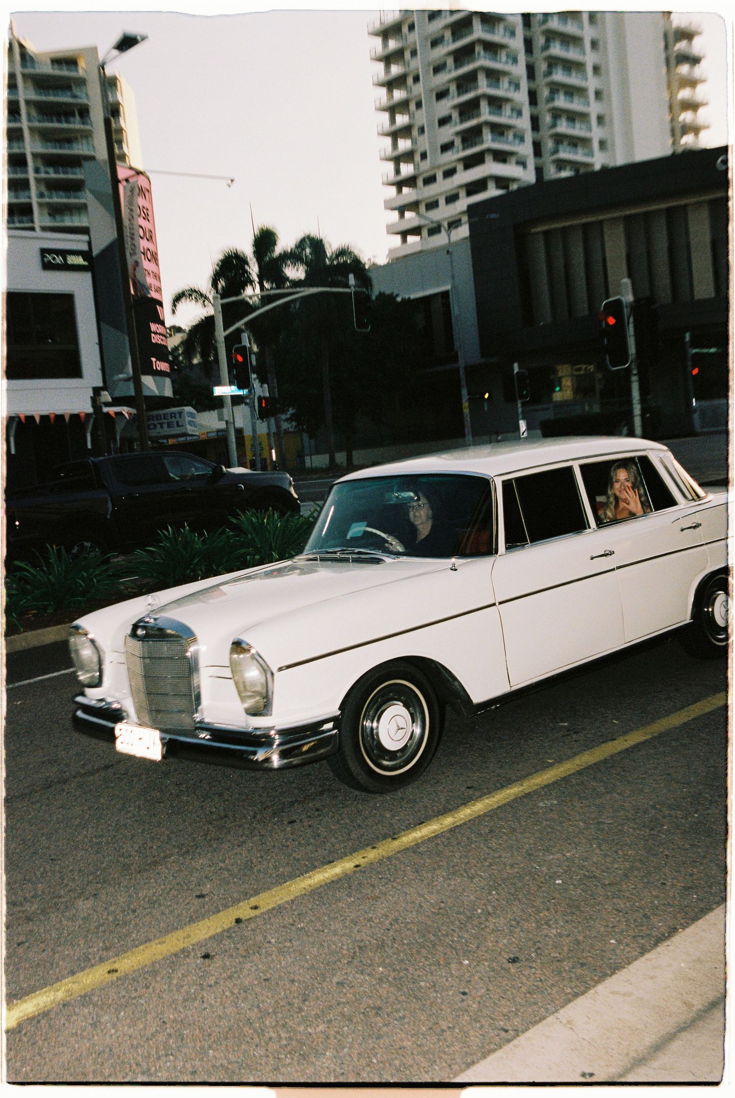 Vintage white Mercedes-Benz wedding car driving through a busy urban intersection with palm trees.