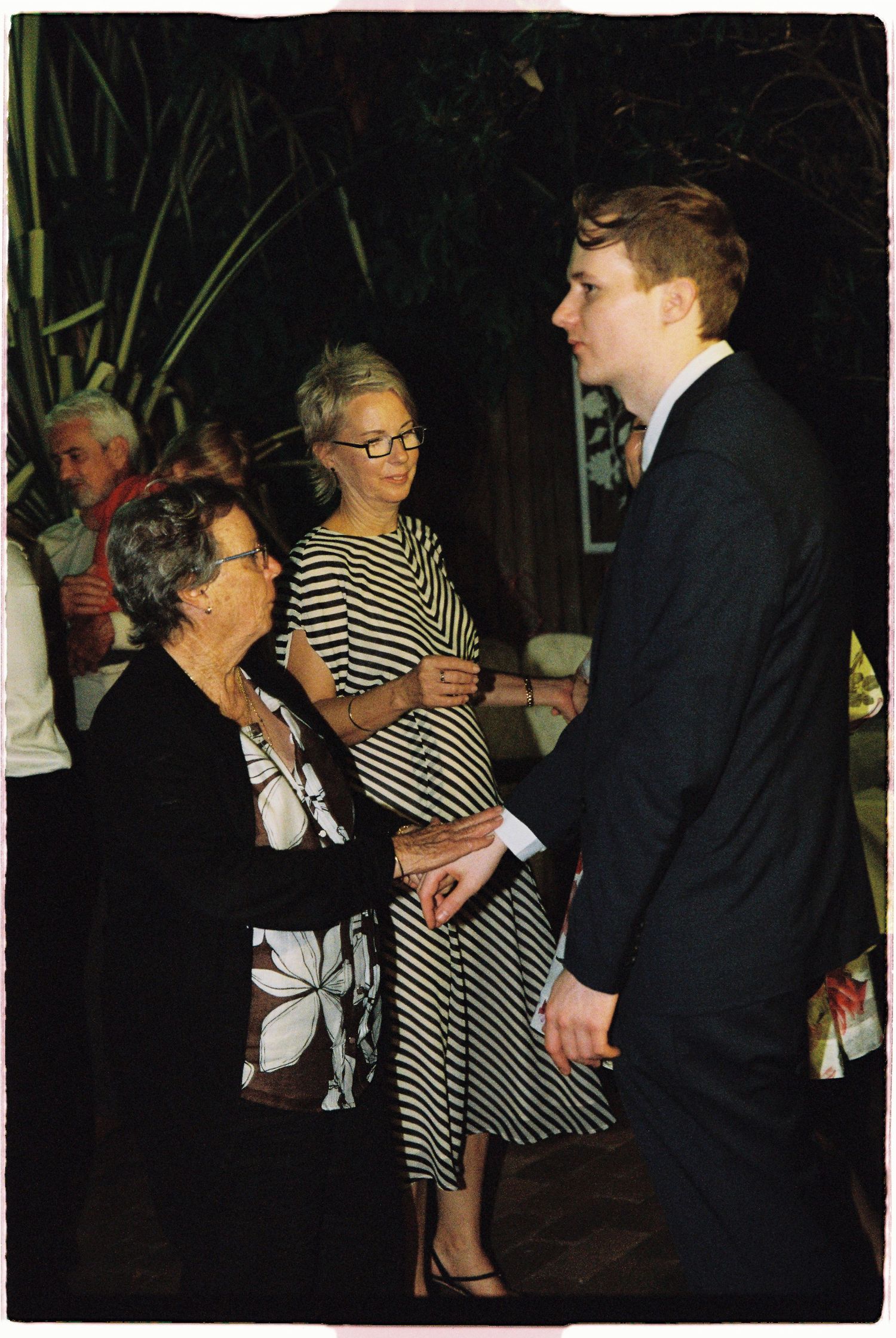 Elderly woman conversing with a young man in a suit and a middle-aged woman in striped dress at a formal gathering.