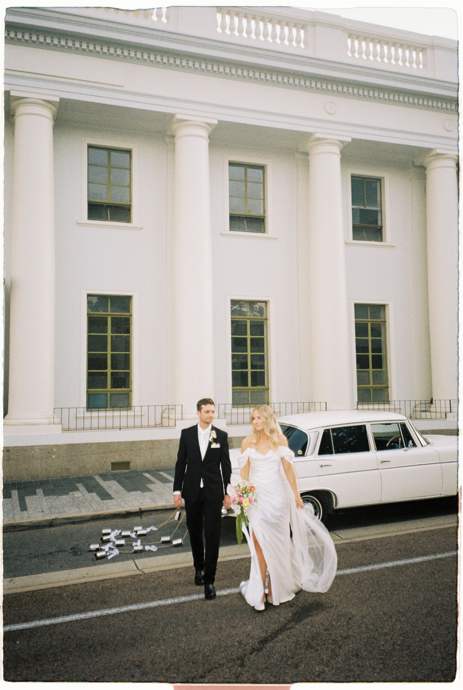 Elegant bride and groom walk past white classic car in front of grand white columned building.