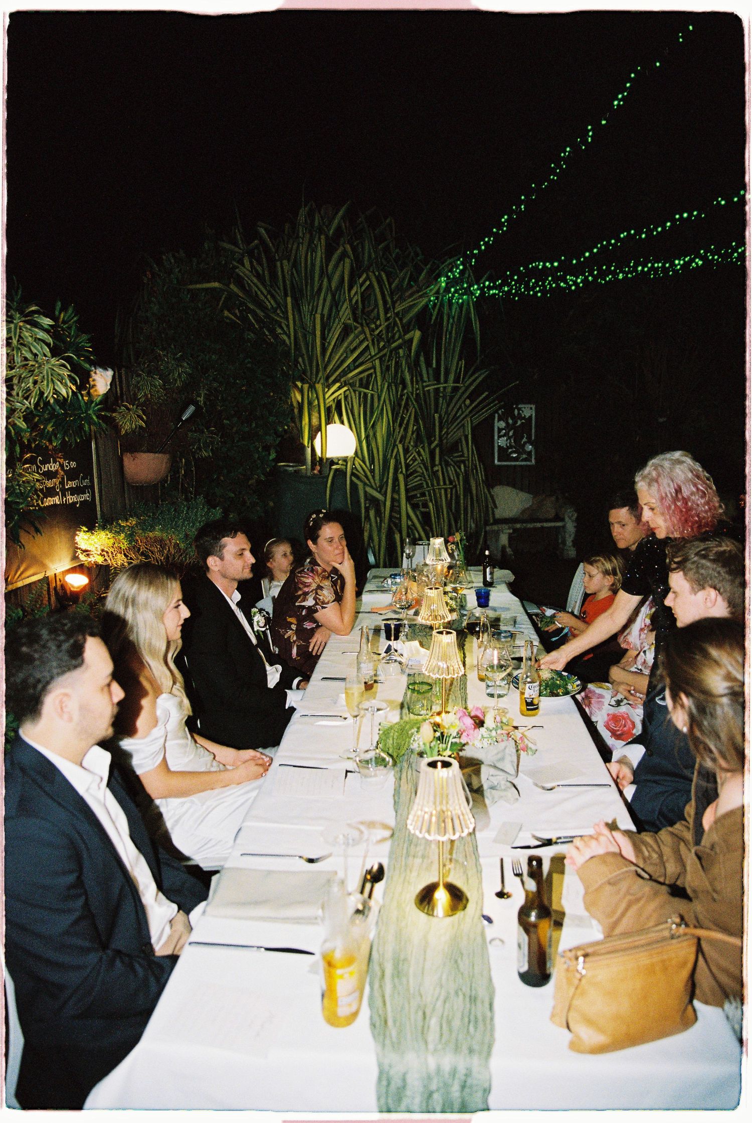 Guests seated at a long elegant outdoor dinner table at night with fairy lights.