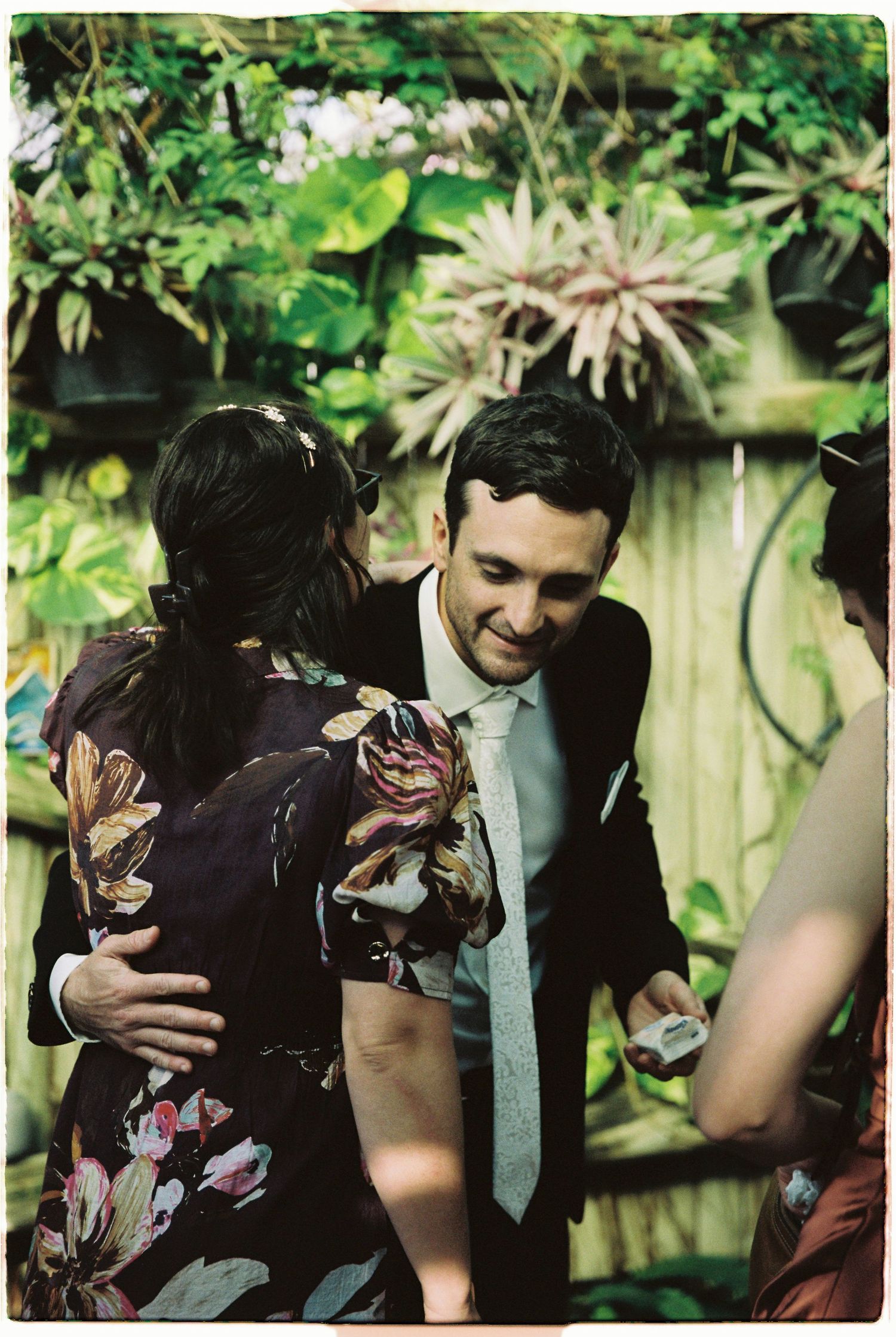 Groom in black suit smiling and embracing wedding guest in floral dress at garden reception.