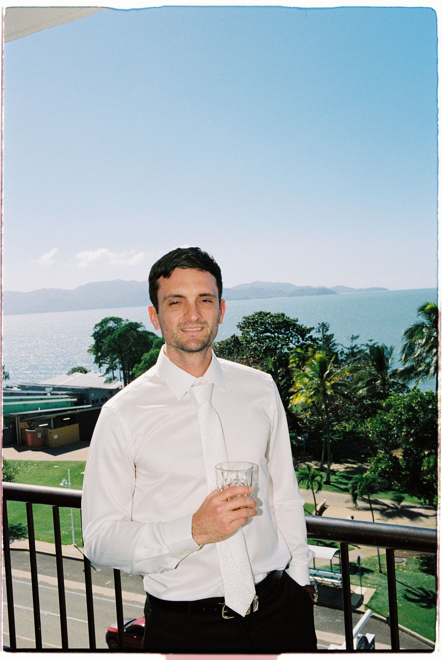 Man in white shirt and tie holding a glass on a balcony overlooking tropical ocean and island views.