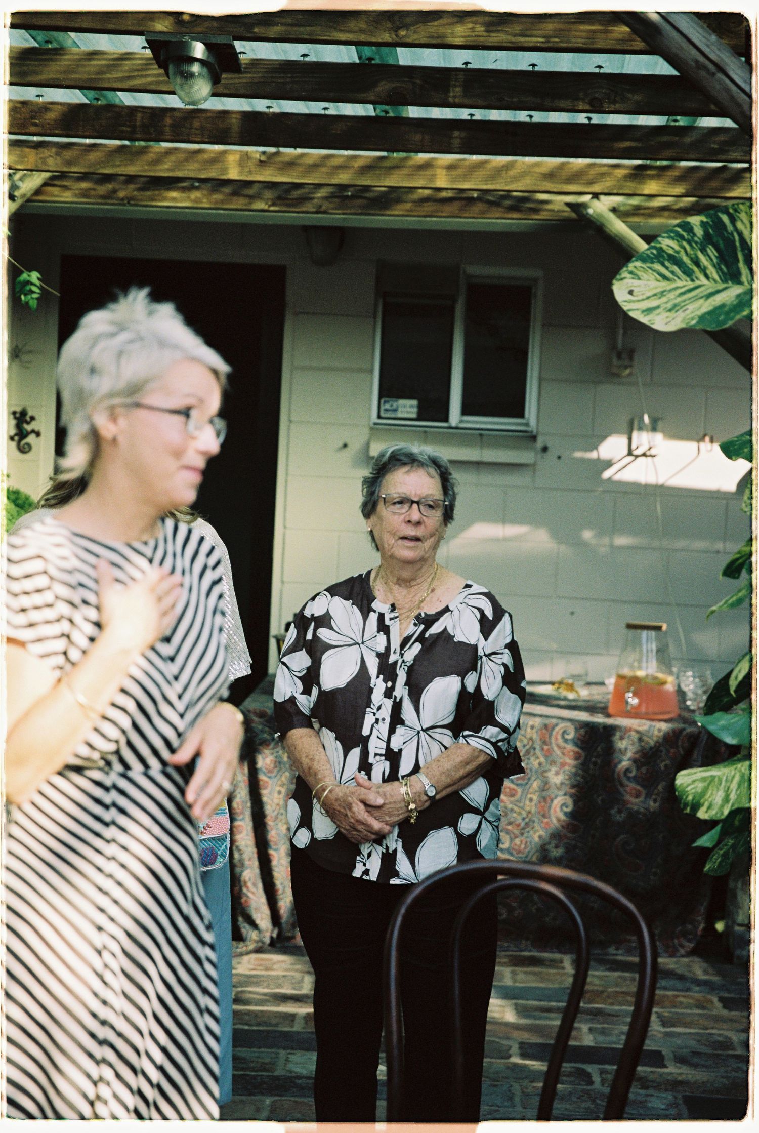 Two older women conversing in sunny backyard patio with tropical plants and rustic decor.