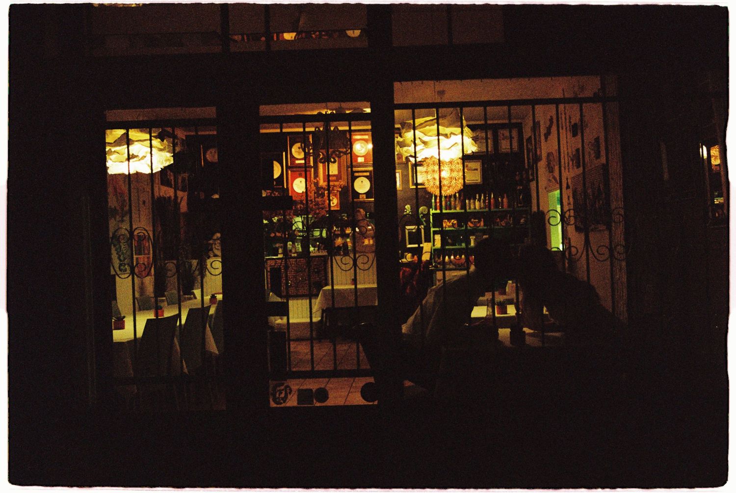 Warmly lit bar or restaurant interior viewed through iron-gated glass doors at night.
