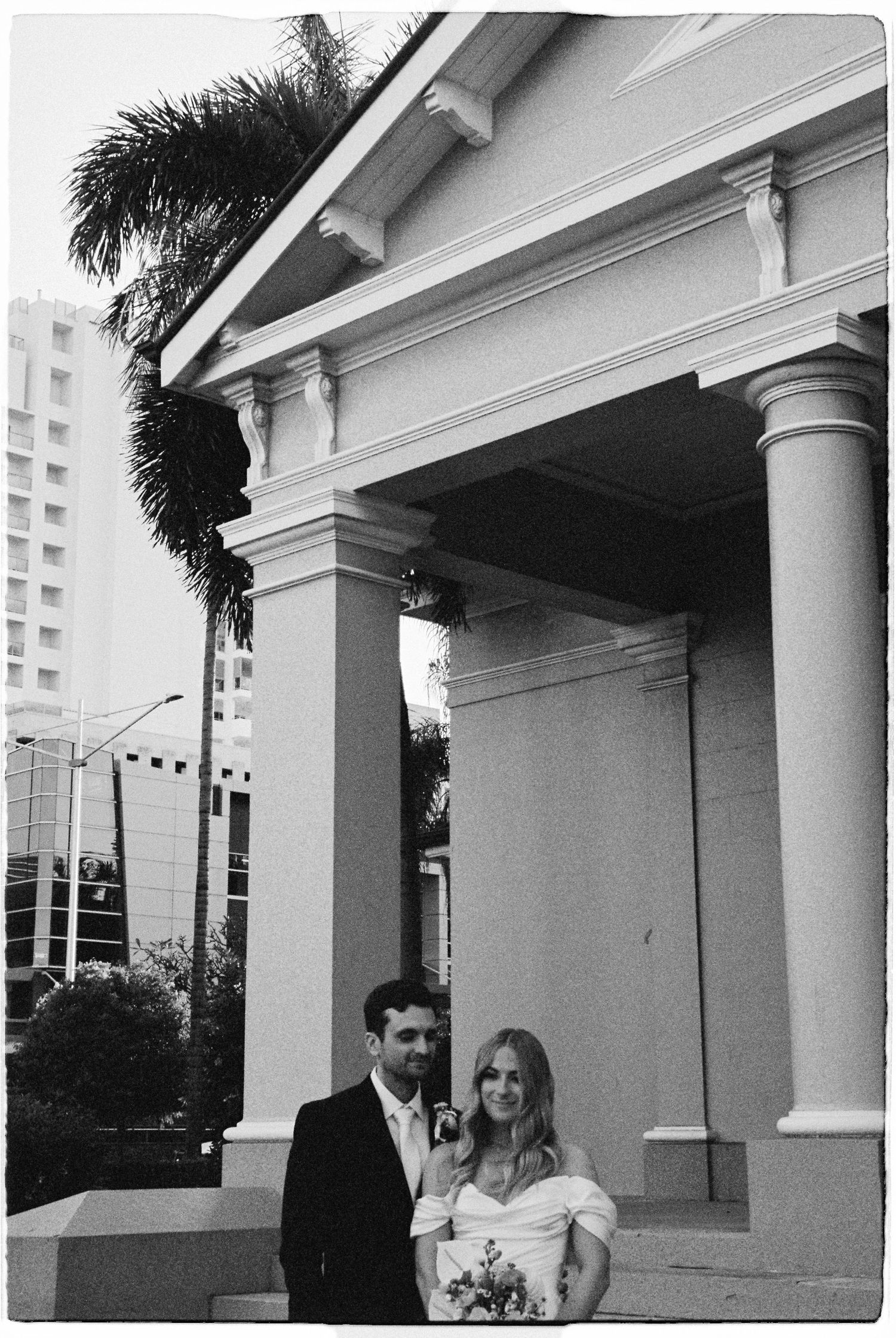 Bride and groom posing outside a neoclassical church with palm trees in black and white photo.