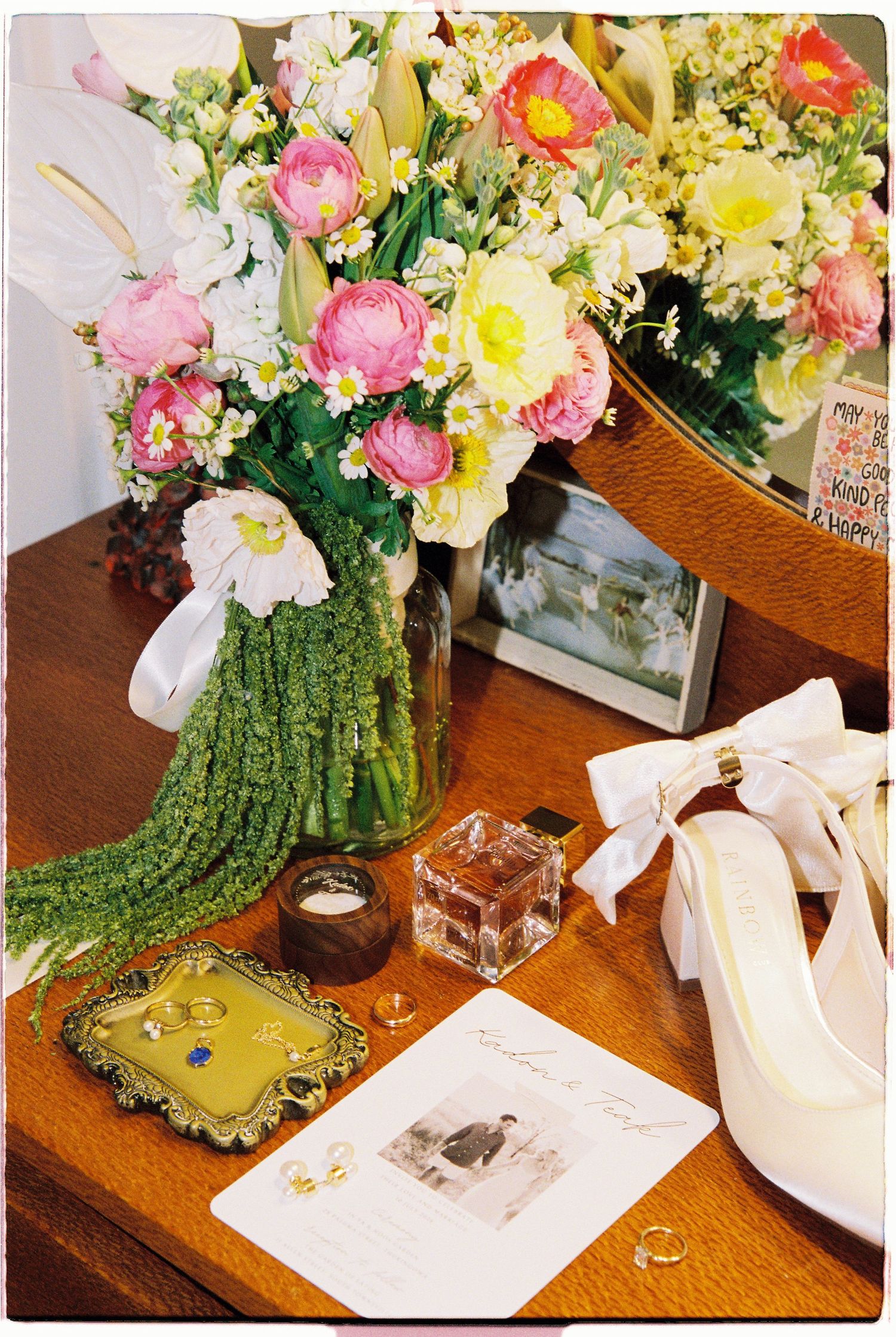 Wedding flat lay with bridal bouquet, white bow heels, rings, and stationery on a wooden table.
