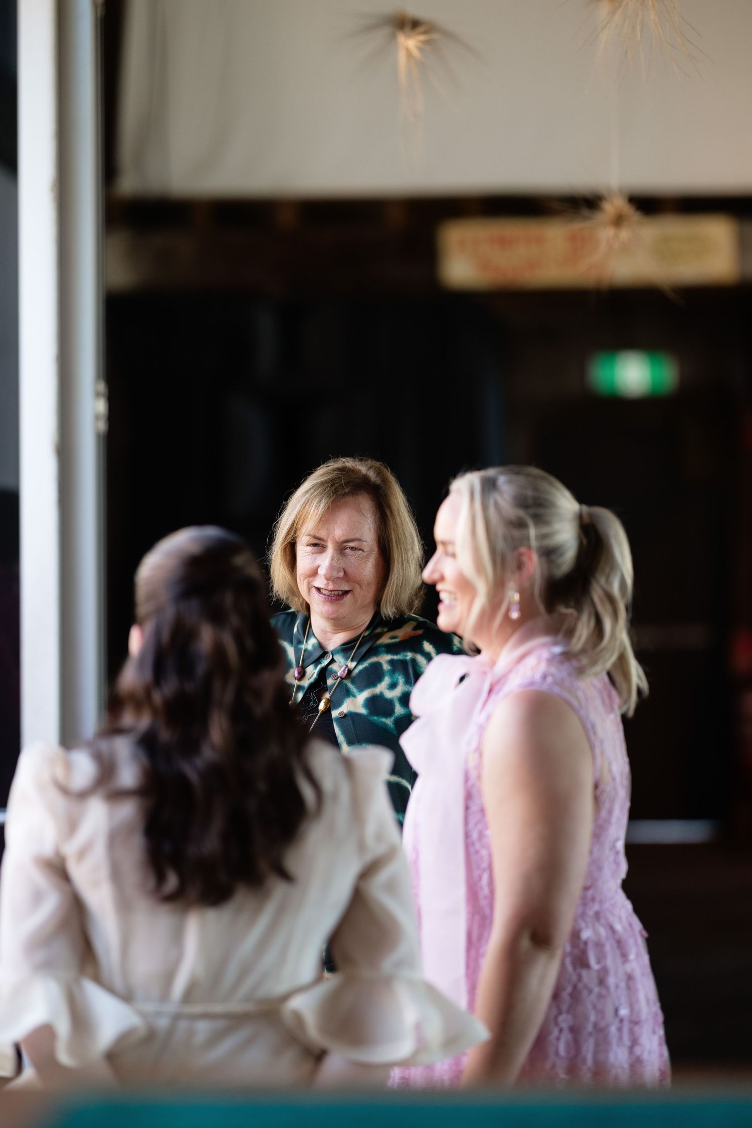 Three women in elegant attire laugh and chat together at a bright and airy wedding reception venue.