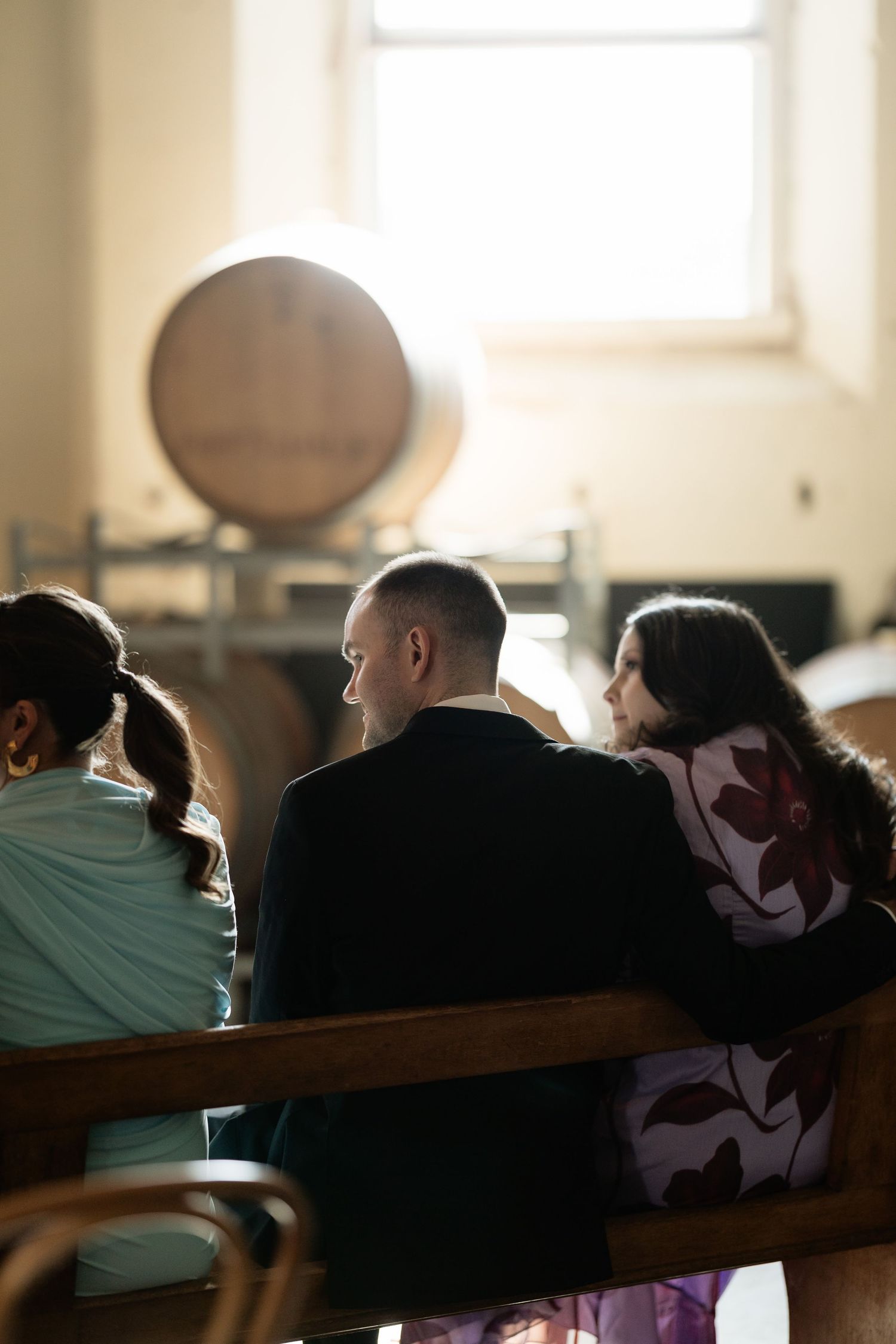 Guests seated in wooden pews at a winery wedding ceremony with oak barrels in the background.