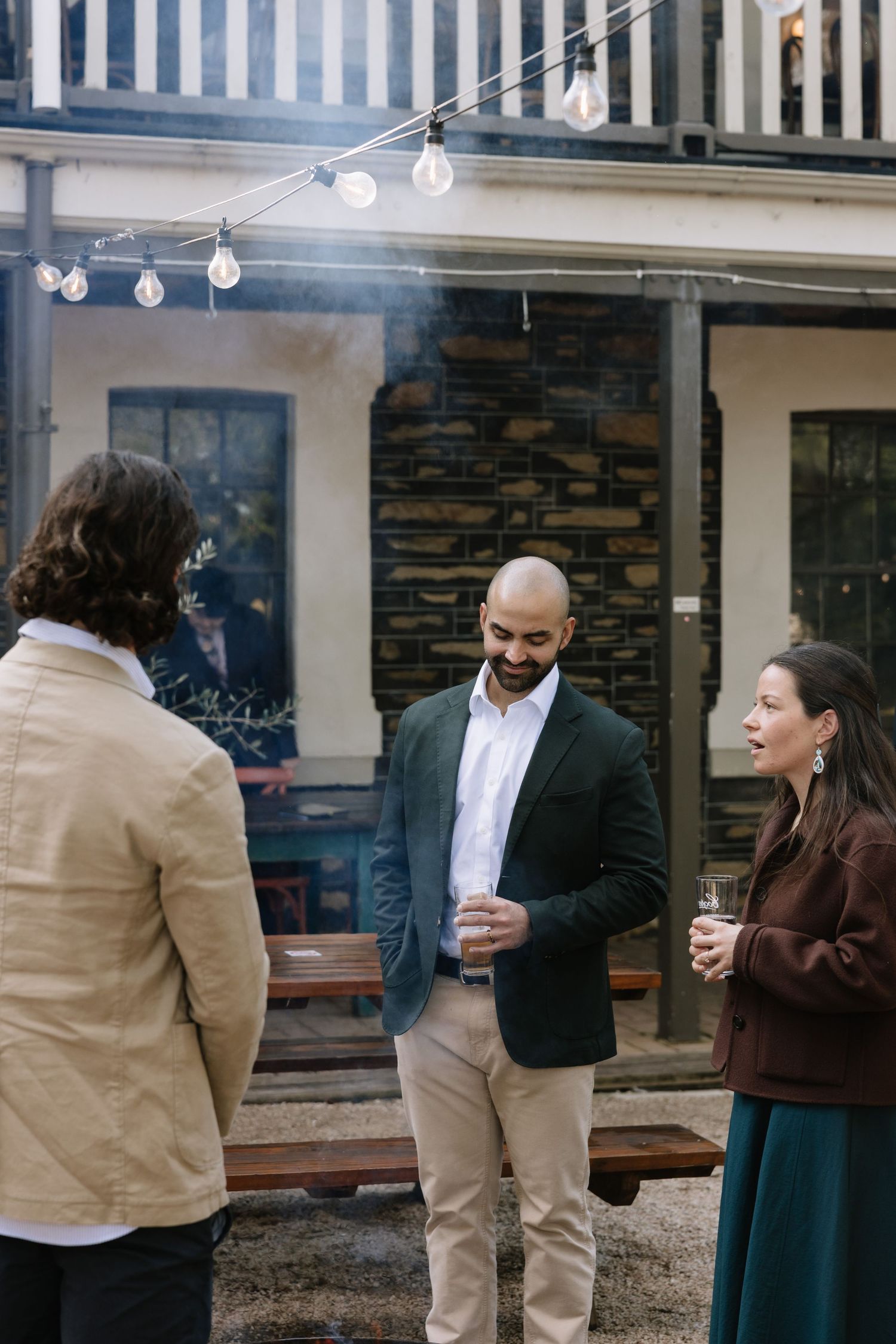 Three people socializing outdoors at a casual gathering, holding drinks near string lights and a stone building.