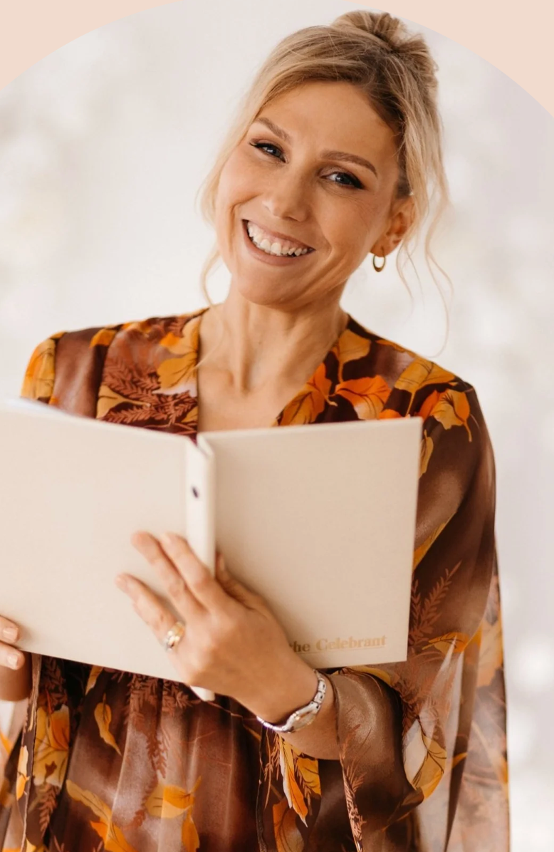 Smiling blonde woman in a brown floral dress holding an open notebook, perfect for wedding celebrant or event planner concepts.