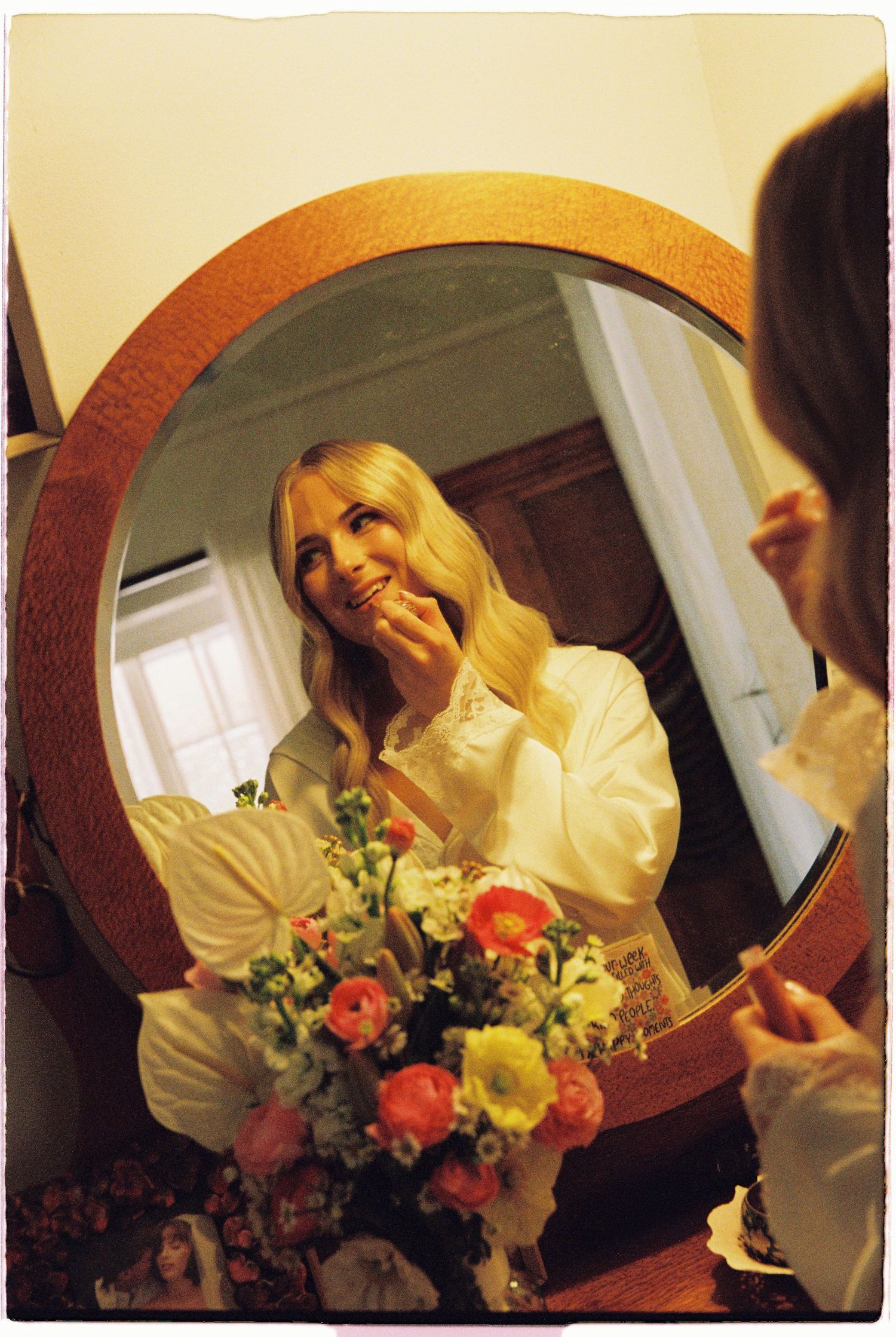 Blonde woman in white blouse smiling at her reflection in a round wooden mirror beside a colorful floral arrangement.