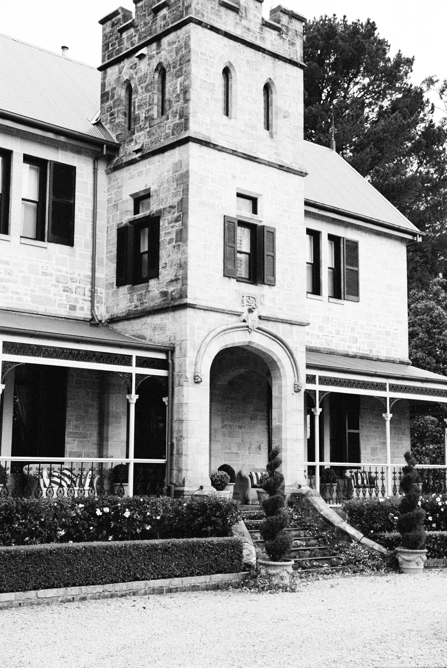 Black and white photo of a historic stone manor house with Gothic tower, arched entrance, shuttered windows, and manicured gardens.