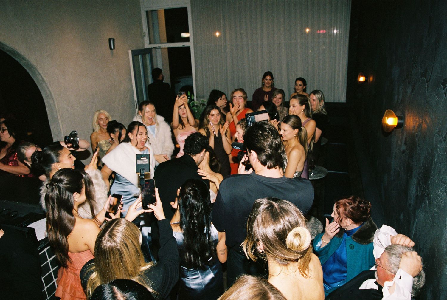 Excited wedding guests surround the bride and groom on the dance floor during a lively reception celebration.