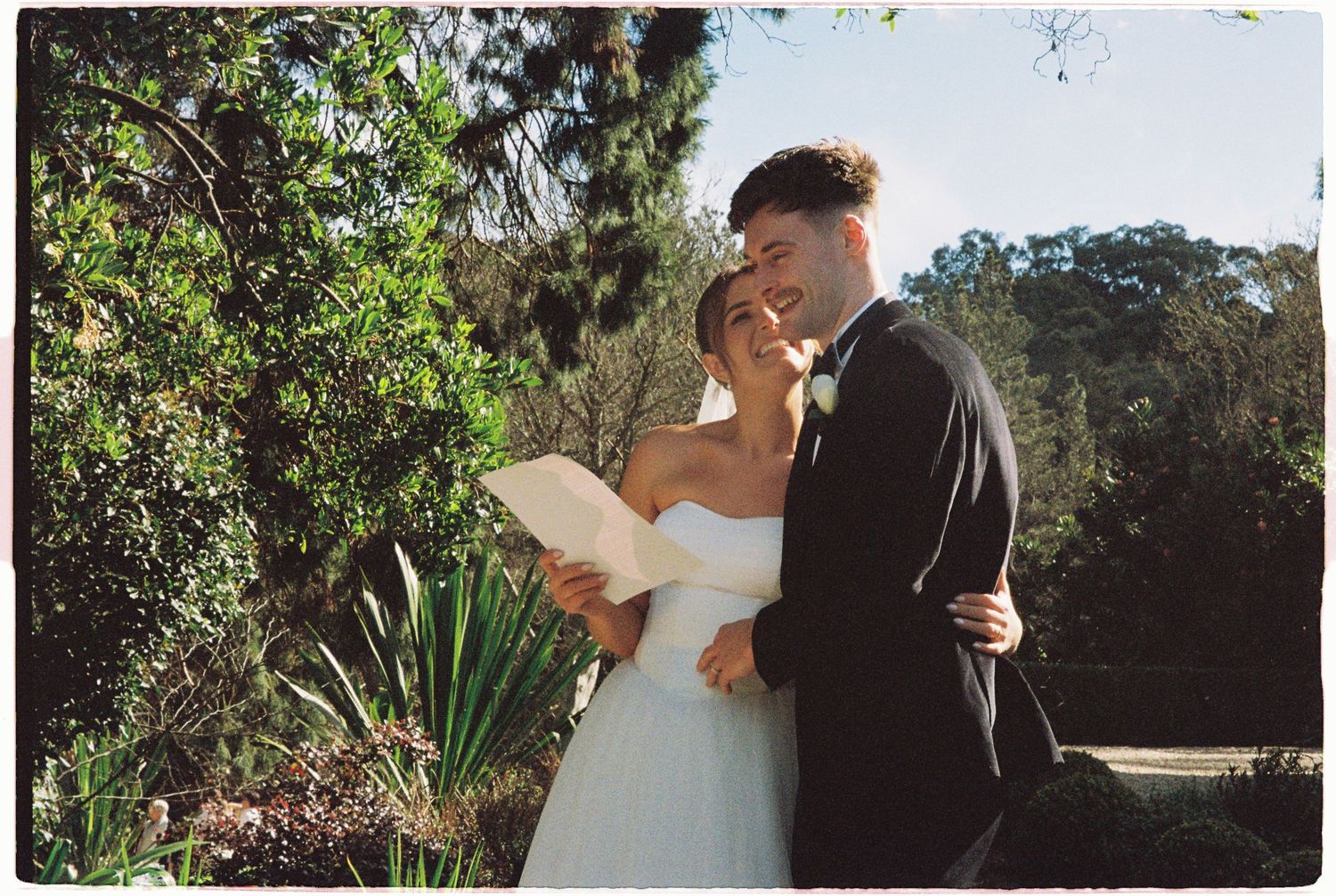 Bride and groom share a tender embrace outdoors surrounded by lush greenery on their wedding day.