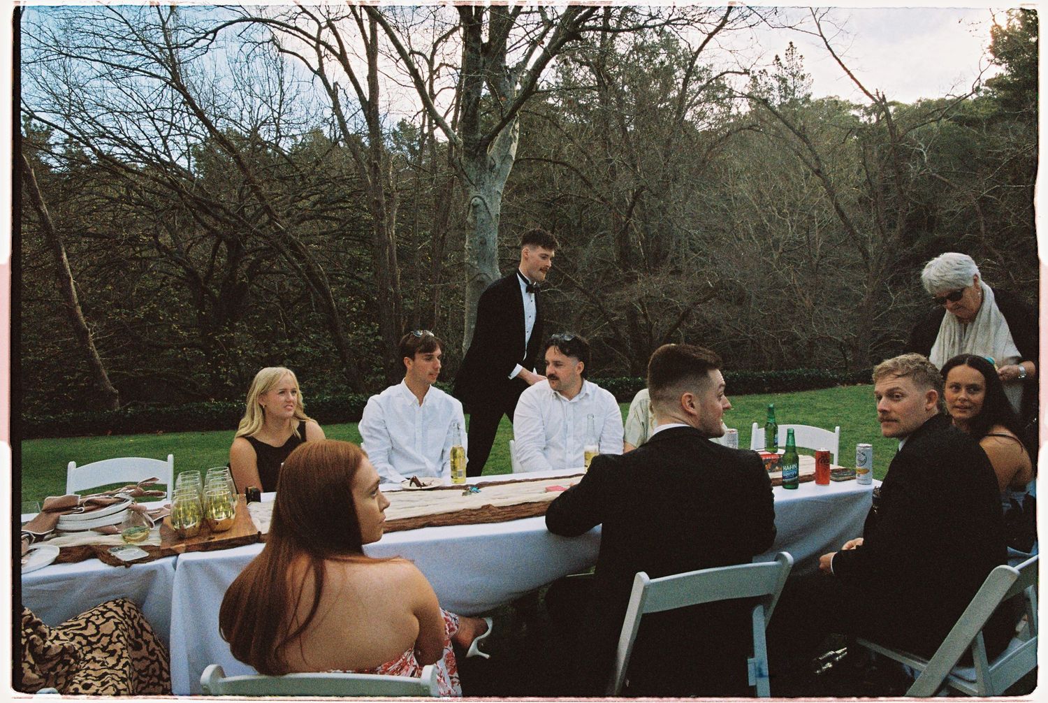 Wedding party guests gathered around long outdoor dining table during al fresco reception in garden setting.
