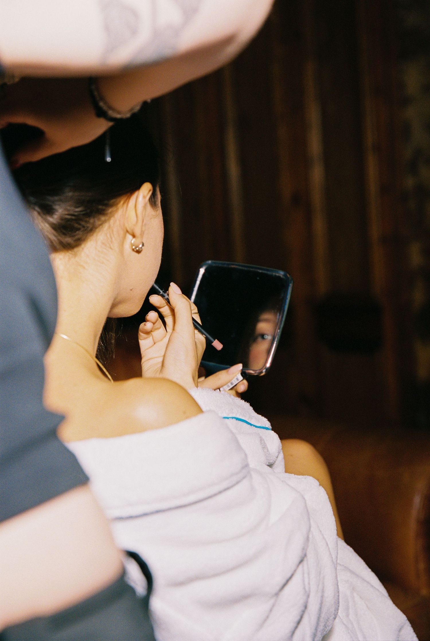 A bride in a white robe checks her earring in a small mirror during wedding preparations.