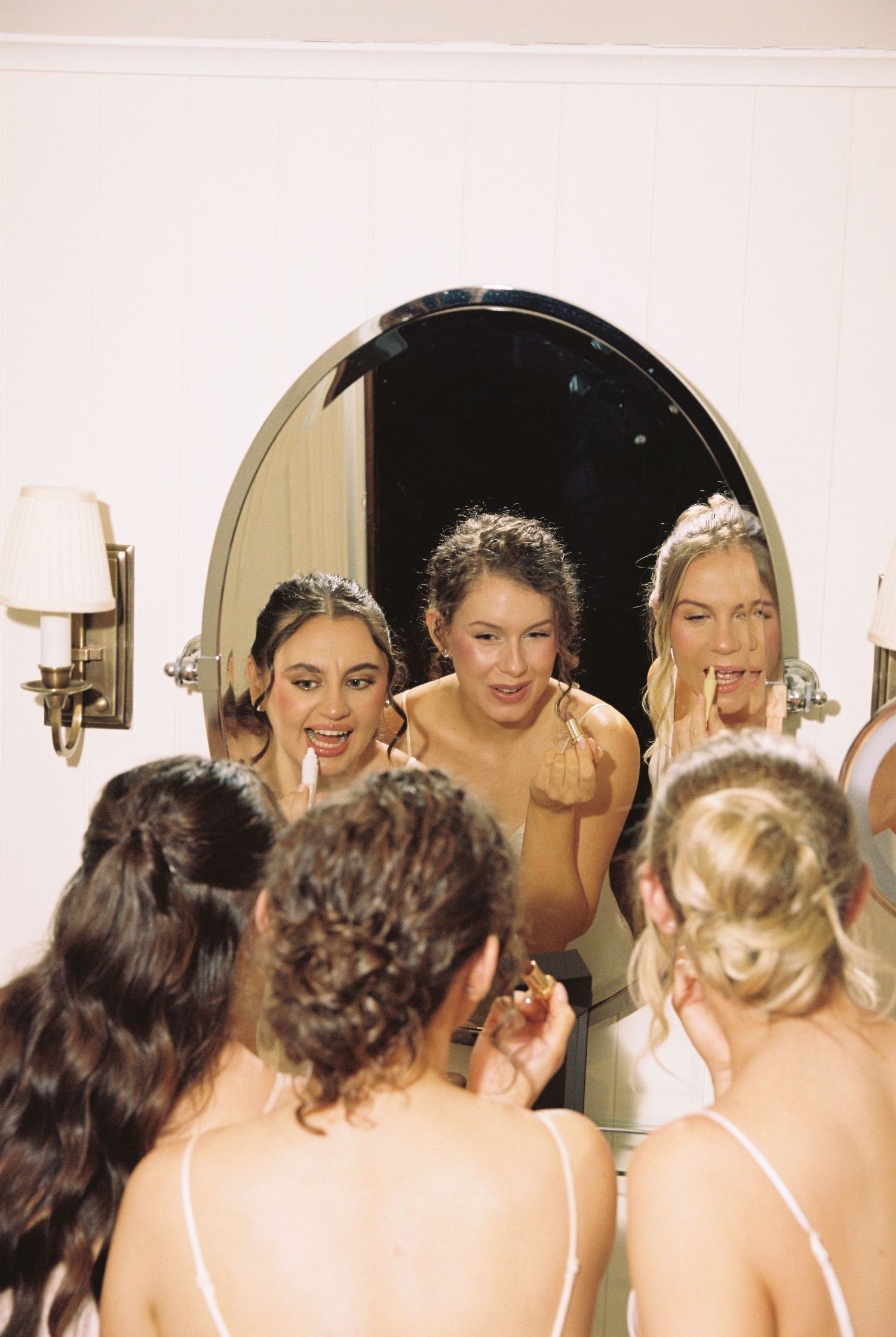 Three bridesmaids in satin dresses applying lipstick while looking into an elegant oval mirror.
