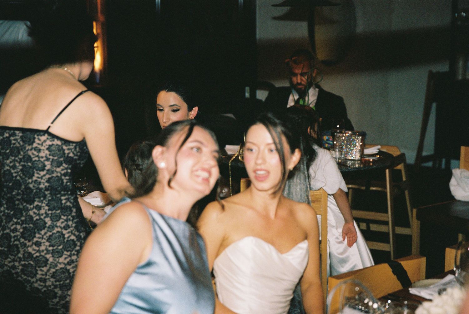 Two women laughing joyfully at a wedding reception dinner table, one in white strapless gown.